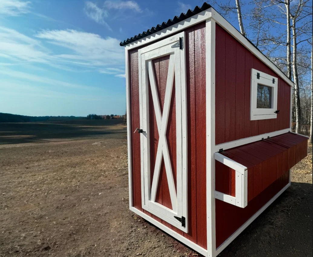 A red barn-style shed with white trim, a sliding door, a small window, and a slanted roof, situated outdoors in a rural area with a wide open field and trees in the background.