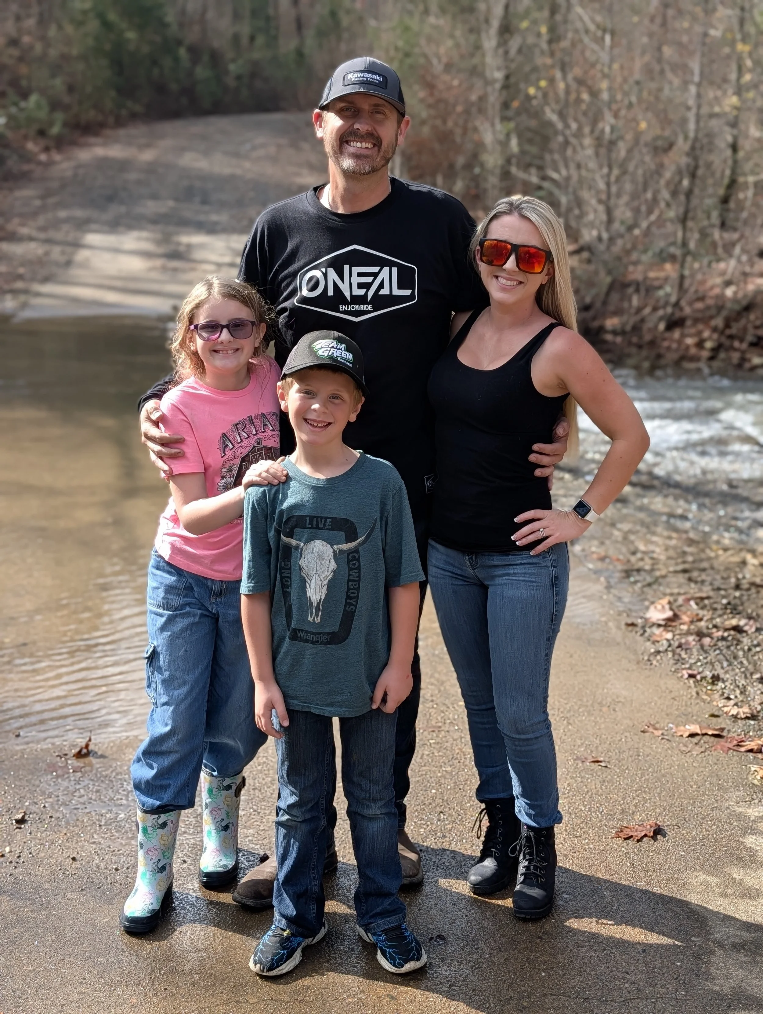 A family of four standing on a riverbank during autumn, smiling for a photo. The father is wearing a black O'NEAL T-shirt and a black cap, the mother in a black tank top and sunglasses, the girl in a pink T-shirt and rain boots, and the boy in a gray T-shirt and cap.
