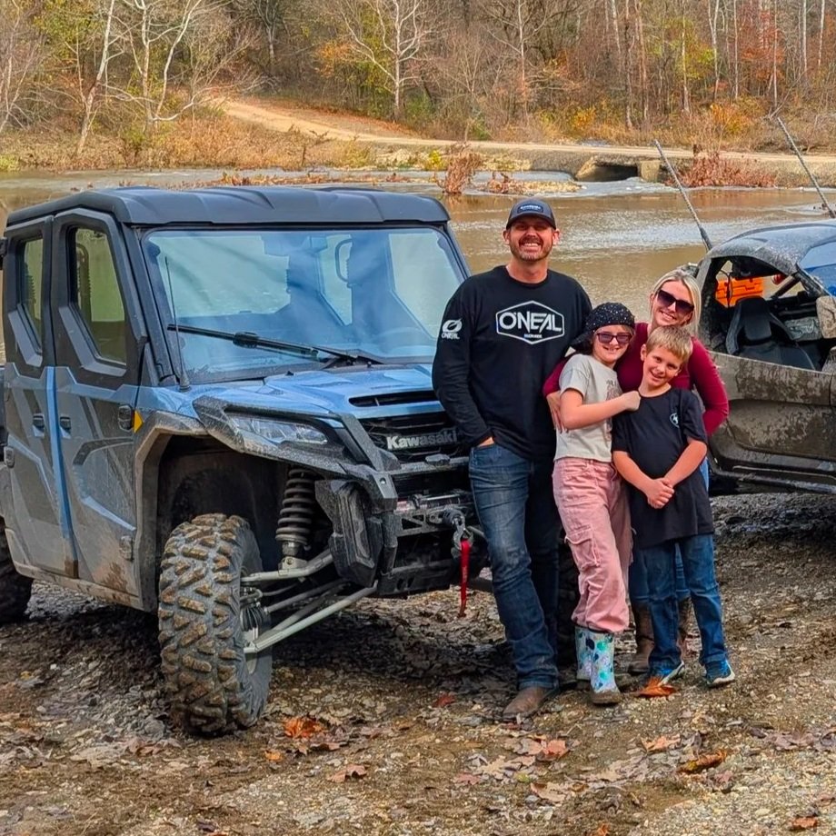 A family of four, with two children, standing beside a blue off-road vehicle and a gray all-terrain vehicle by a river with a wooded background, in autumn.