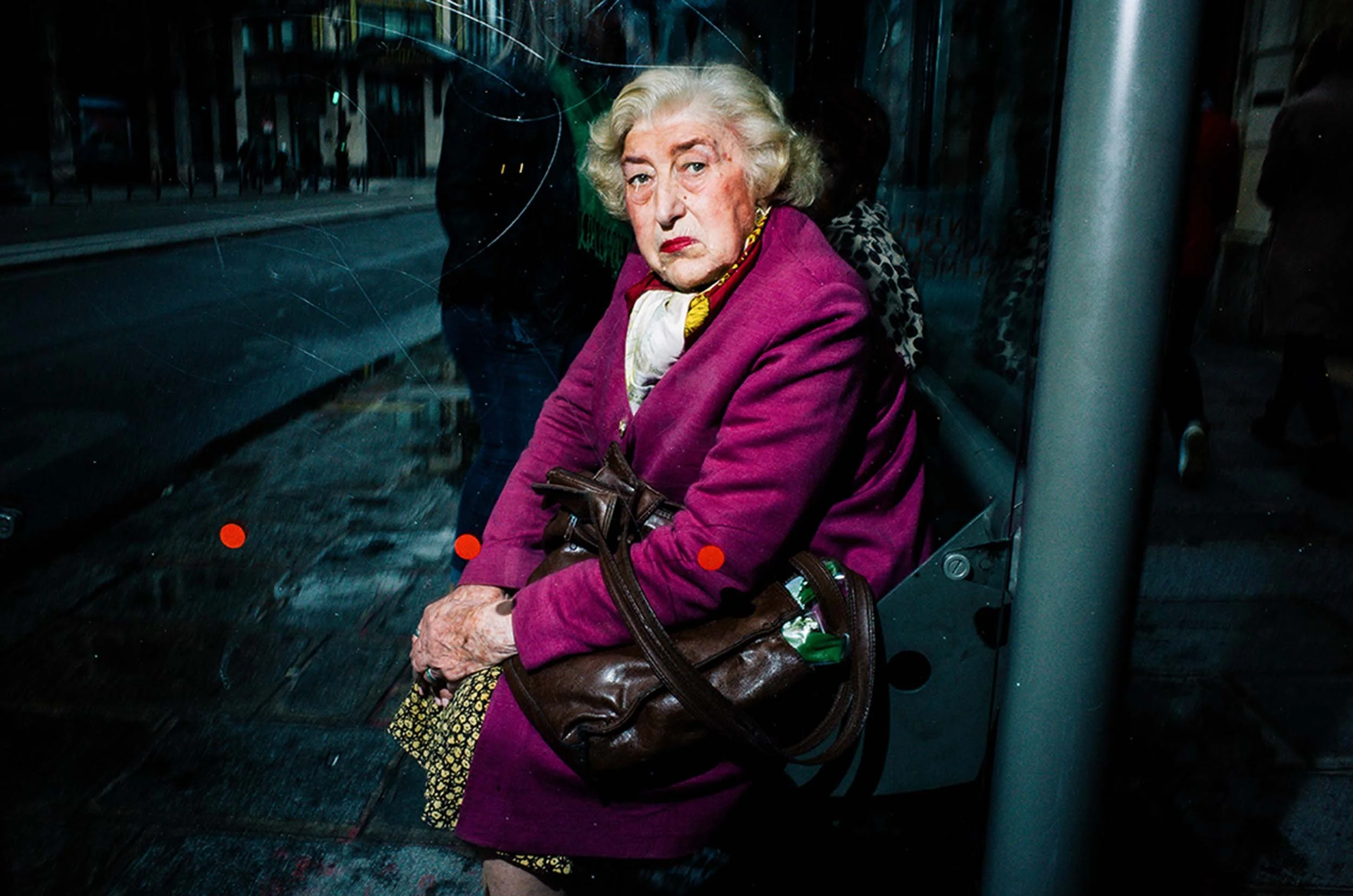 An elderly lady in a purple coat sits and looks directly at the camera at a bus stop.