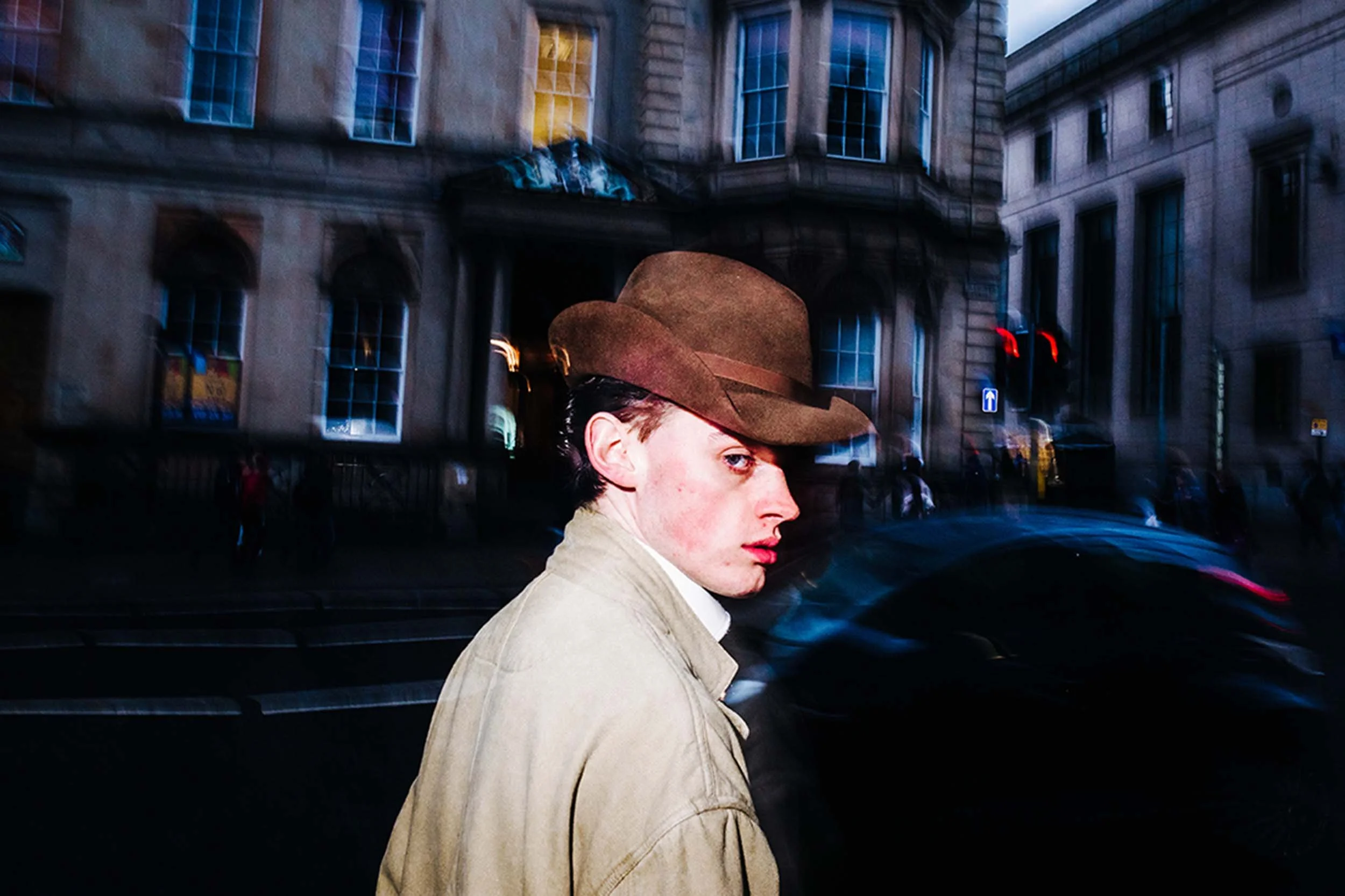 A younger gentleman wearing a trench coat and an old brown hat walking through the city streets in the late afternoon.