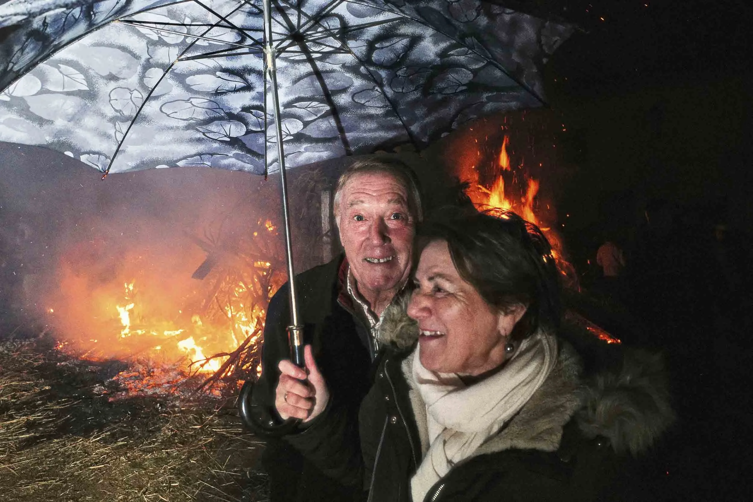A couple underneath an umbrella standing in front of a fire.