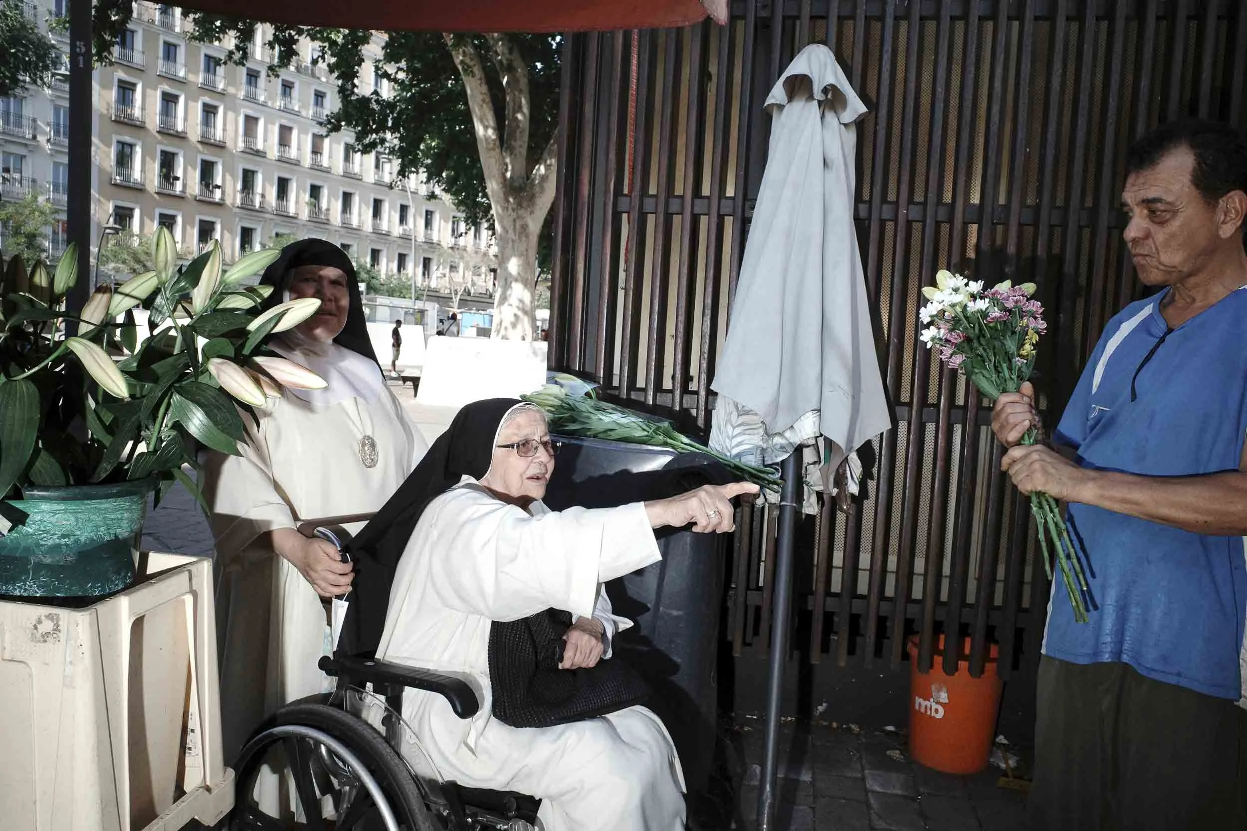A nun pushes a second nun in a wheelchair who is pointing at some flowers that a man is holding.