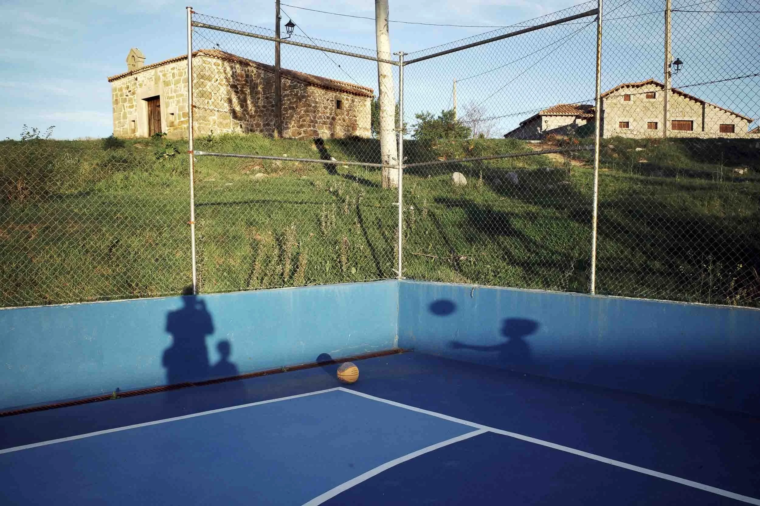 An empty, blue basketball court as the sun sets and you can see the shadow of the photographer and a kid playing.