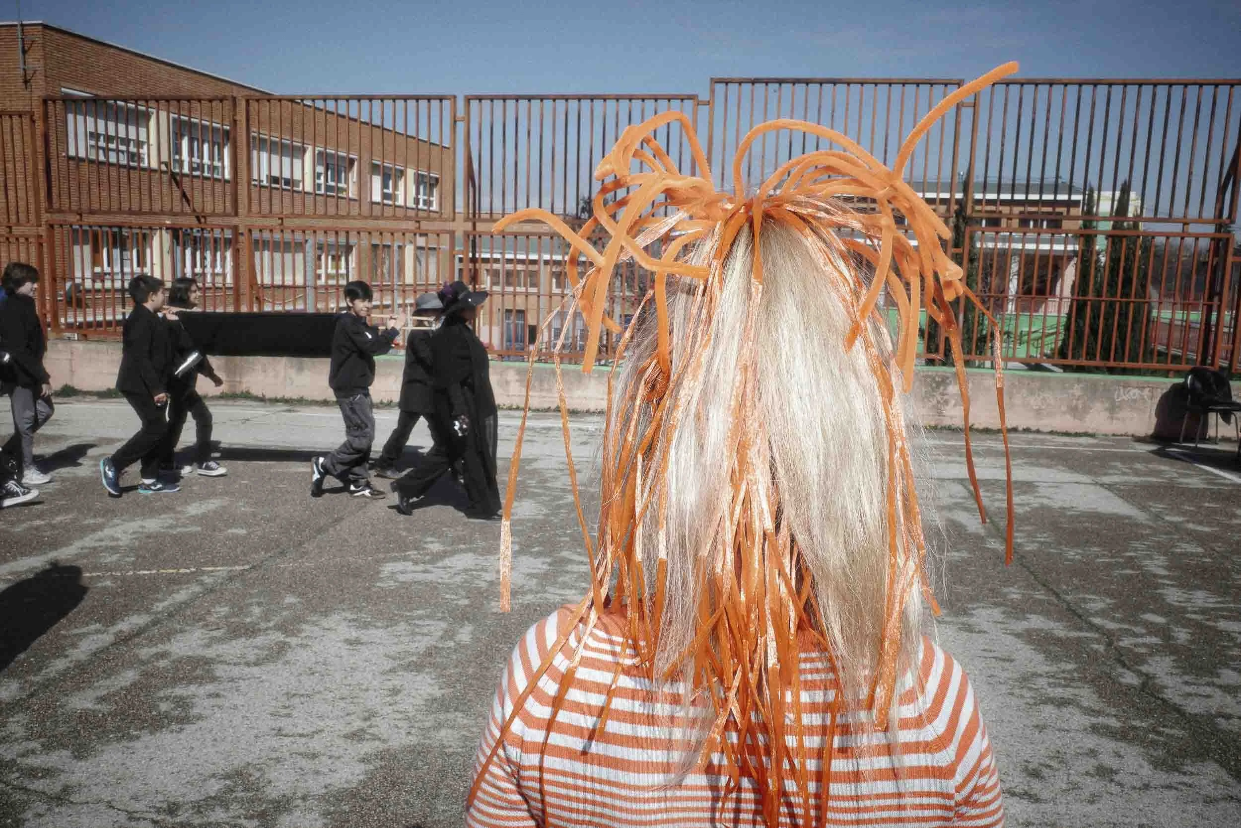 A young girl with orange things in her hair faces away from the camera while some schoolboys walk in the background.