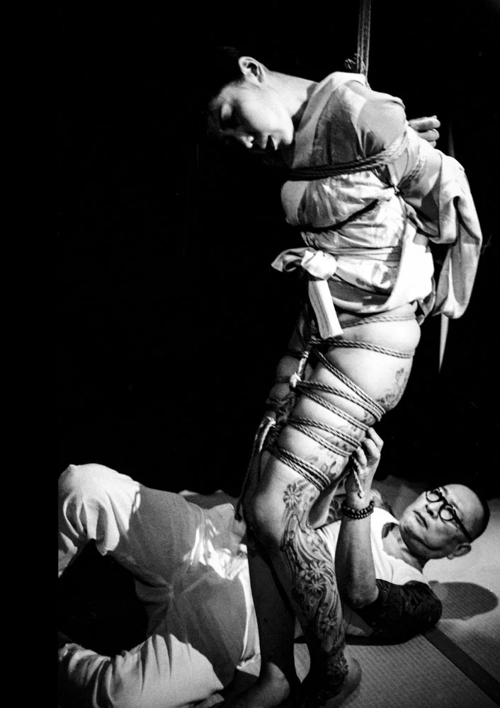Black and white photo of a Japanese male being tied in shibari by another Japanese man who is laying on the ground.