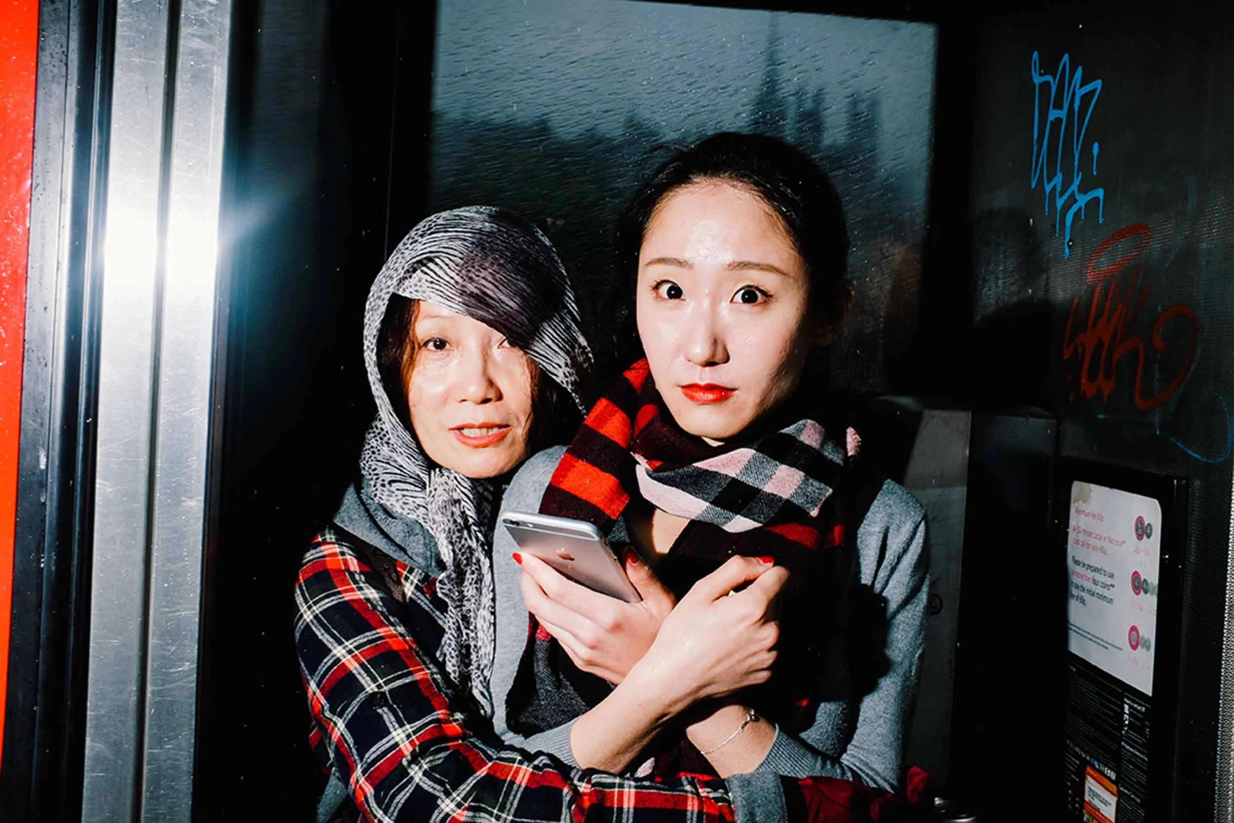 Two asian ladies sheltered inside a phone booth and one holding a mobile phone.