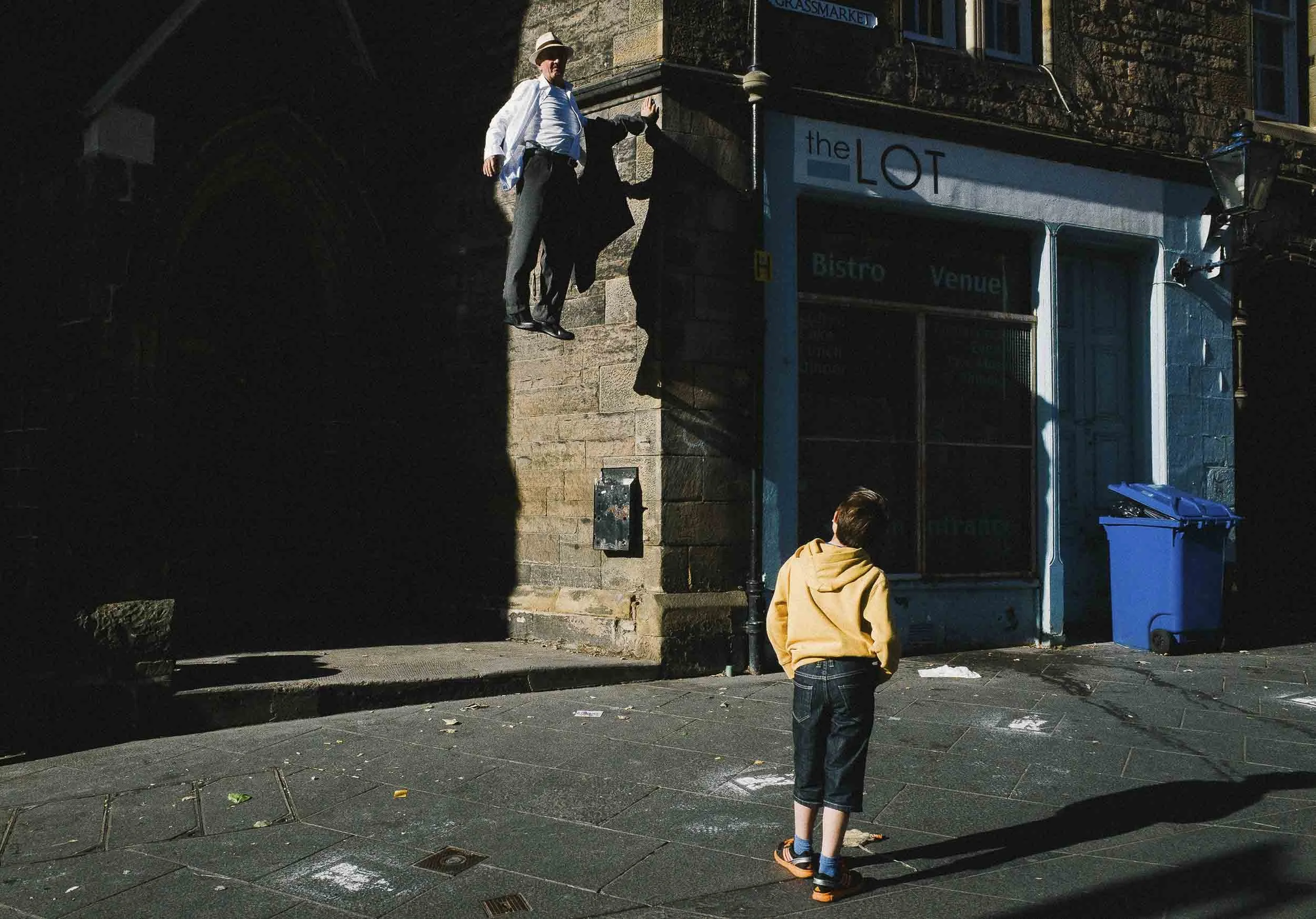 A man is somehow suspended in the air next to a building while a young boy watches on.