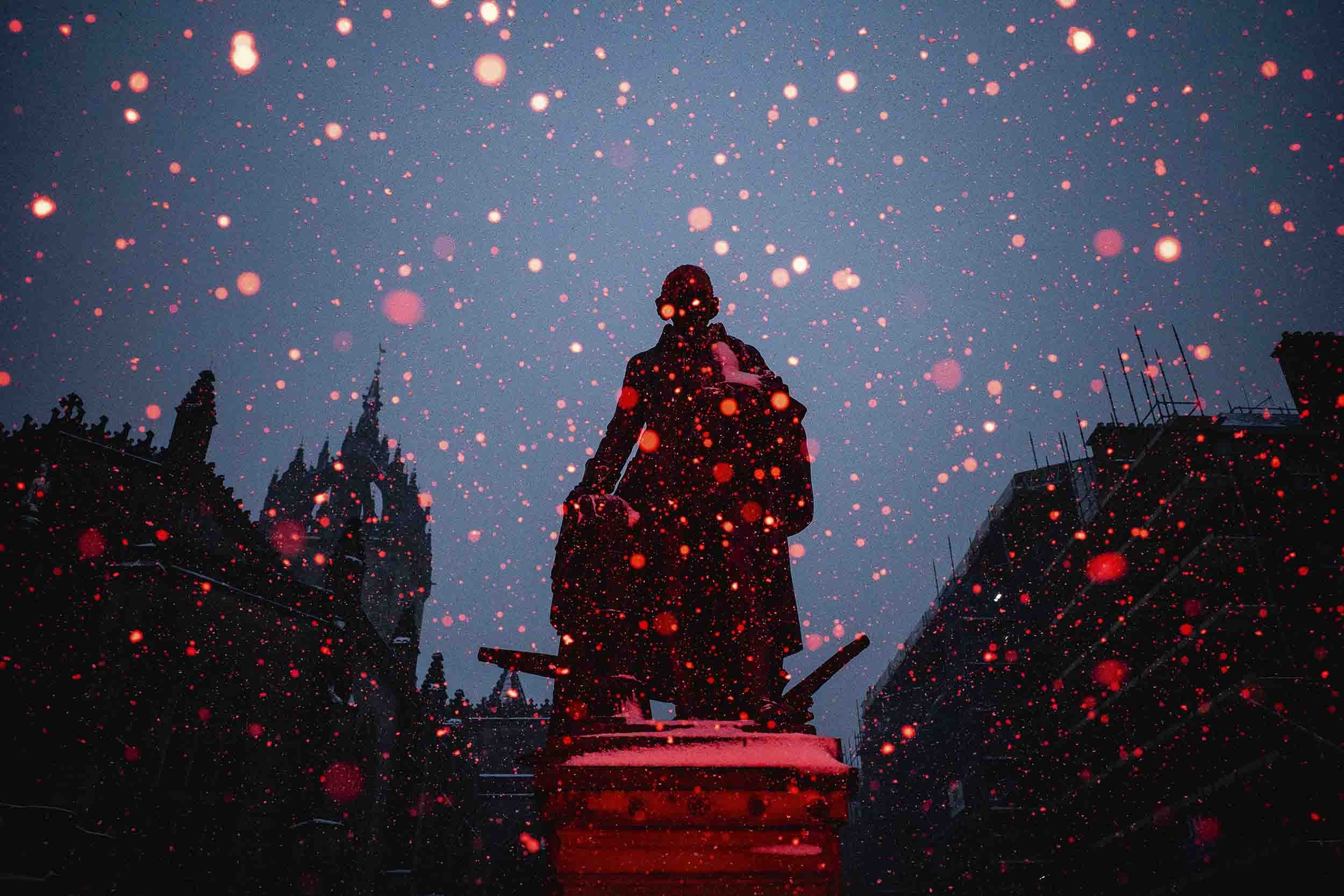 A statue and snow drops bathed in red light sit in front of a blue sky in the evening.