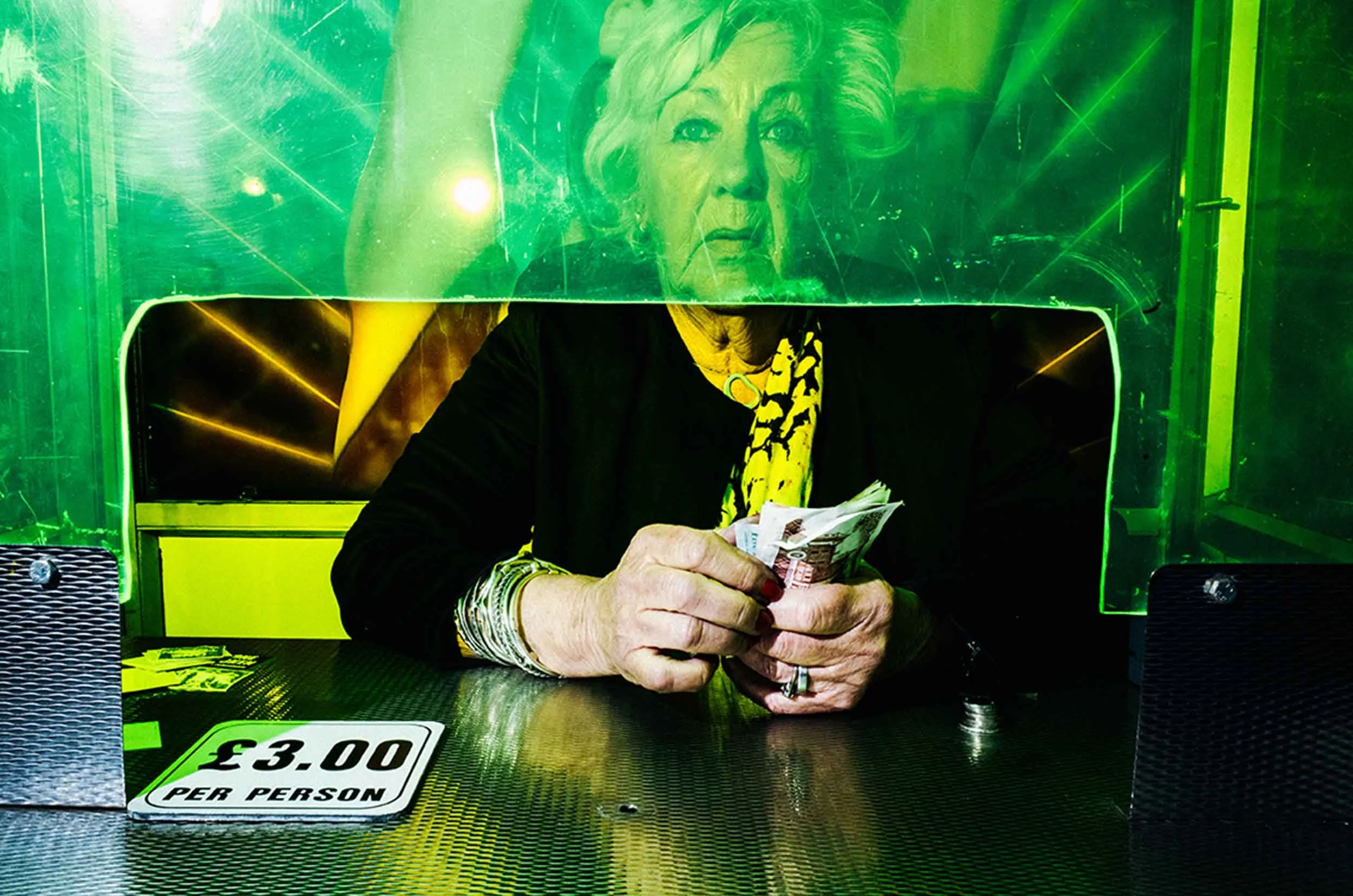 Photo of an elderly lady sitting inside a ticketing booth behind green perspex.