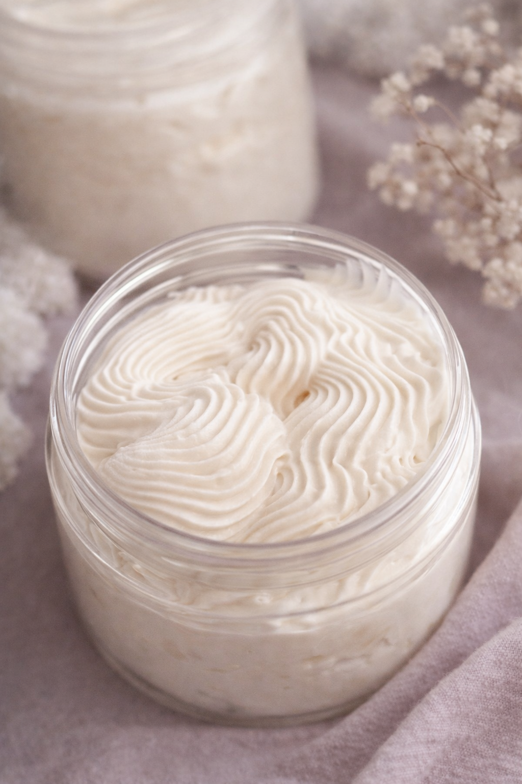 Open jar of white cream with decorative swirl pattern, placed on a soft pink cloth with white flowers and another jar in the background.