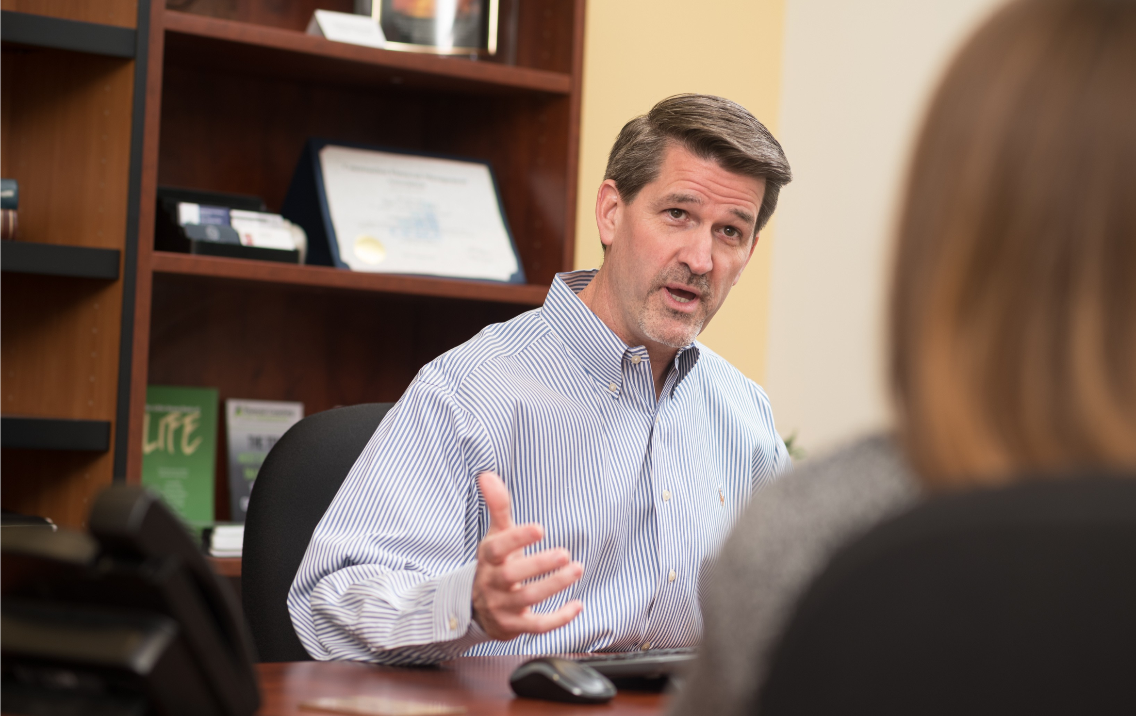 A middle-aged man in a striped shirt having a conversation with a woman in an office setting, gesturing with his hand.