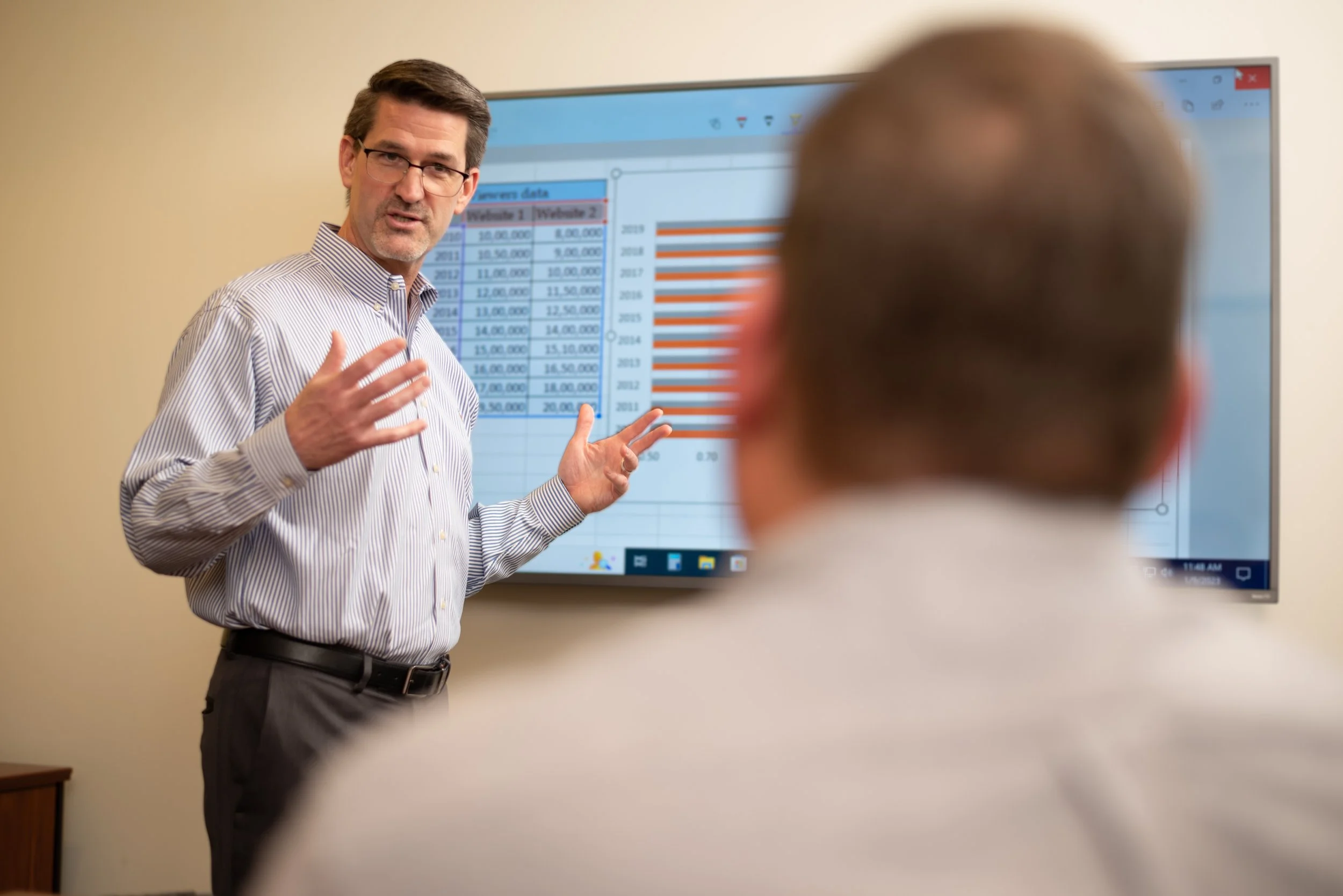 A man with glasses and a striped shirt is giving a presentation in front of a large screen displaying data and bar graphs, while two people are listening.
