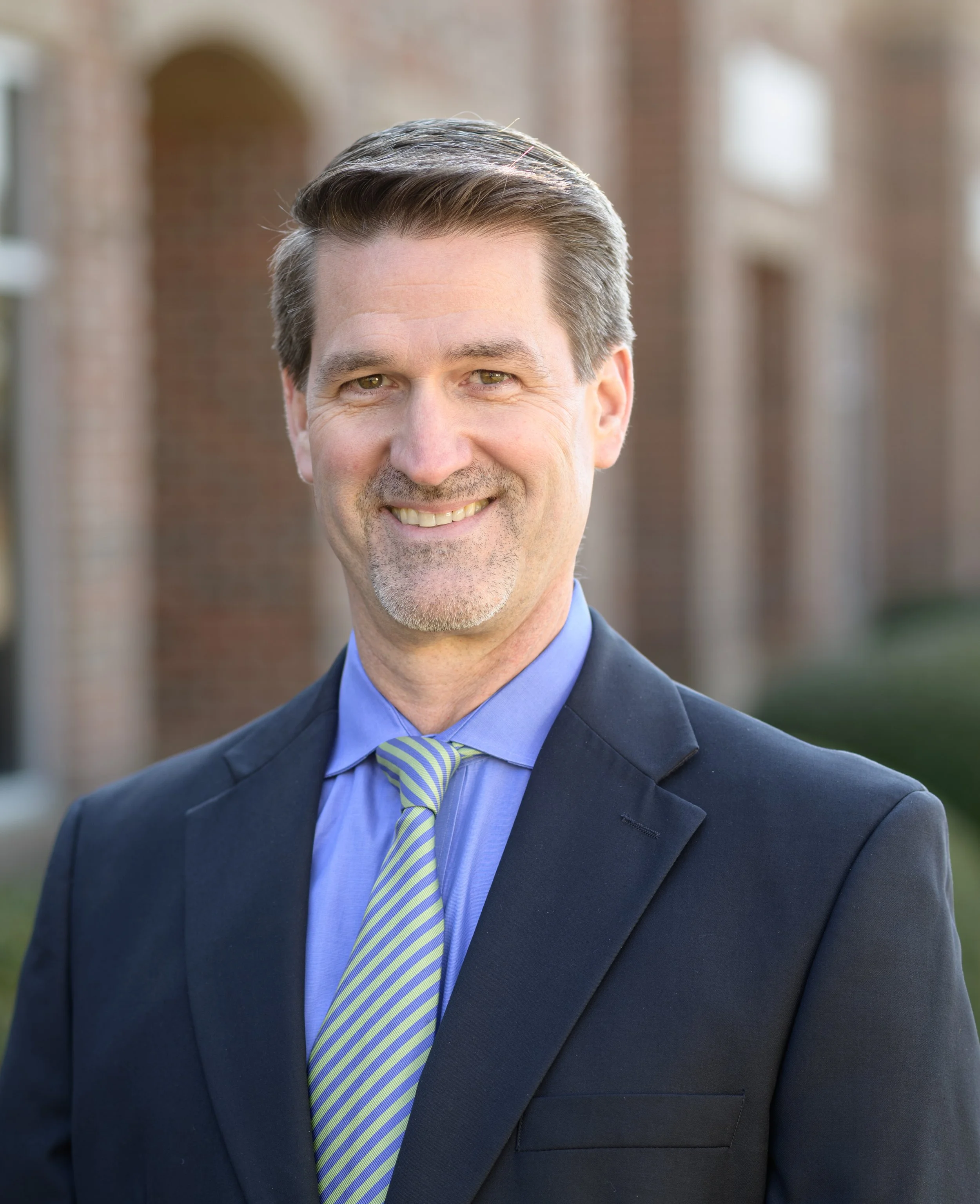 A smiling middle-aged man wearing a dark suit, blue shirt, and green-striped tie, standing outdoors in front of a brick building.