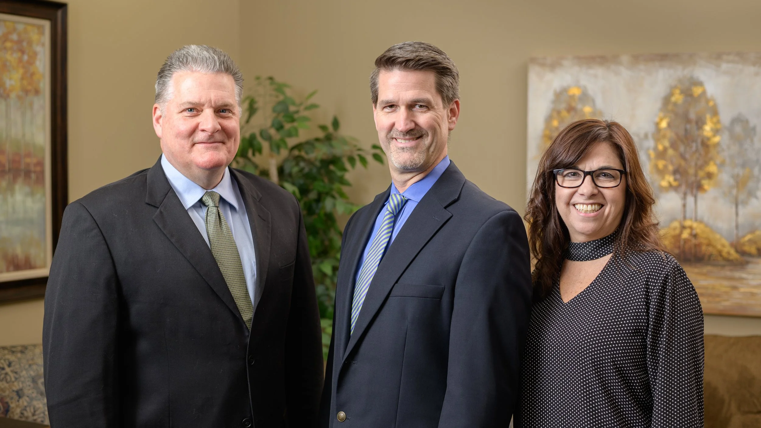 Three smiling professionals in suits and business attire standing in an office or conference room with artwork and plants in the background.