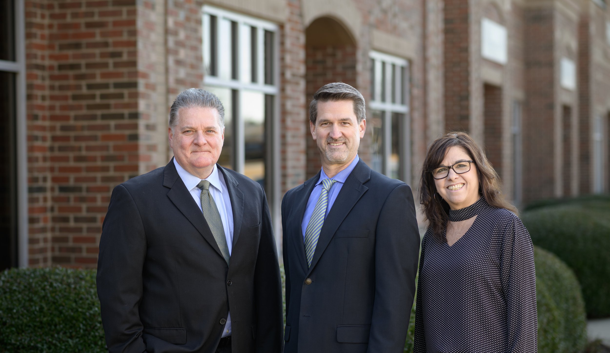 Three professionally dressed individuals standing outside in front of a brick building, smiling at the camera.