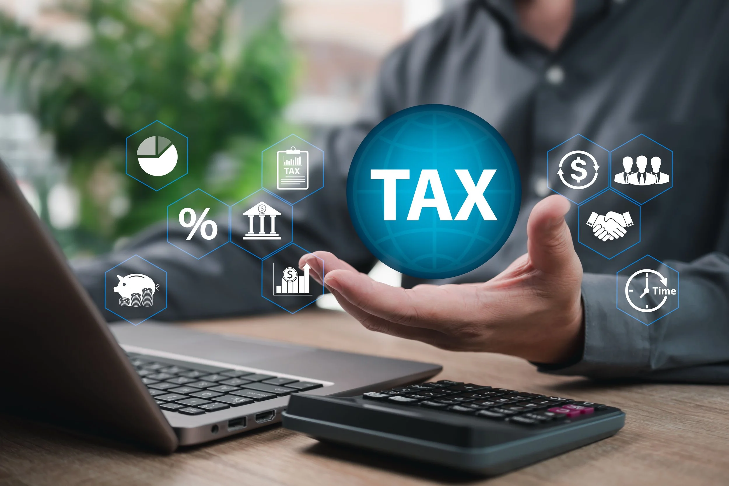 A person holding a digital globe with the word 'TAX' in the center, surrounded by icons representing financial concepts like percentage, government building, bar chart, piggy bank, and clock, with a laptop, calculator, and greenery in the background.