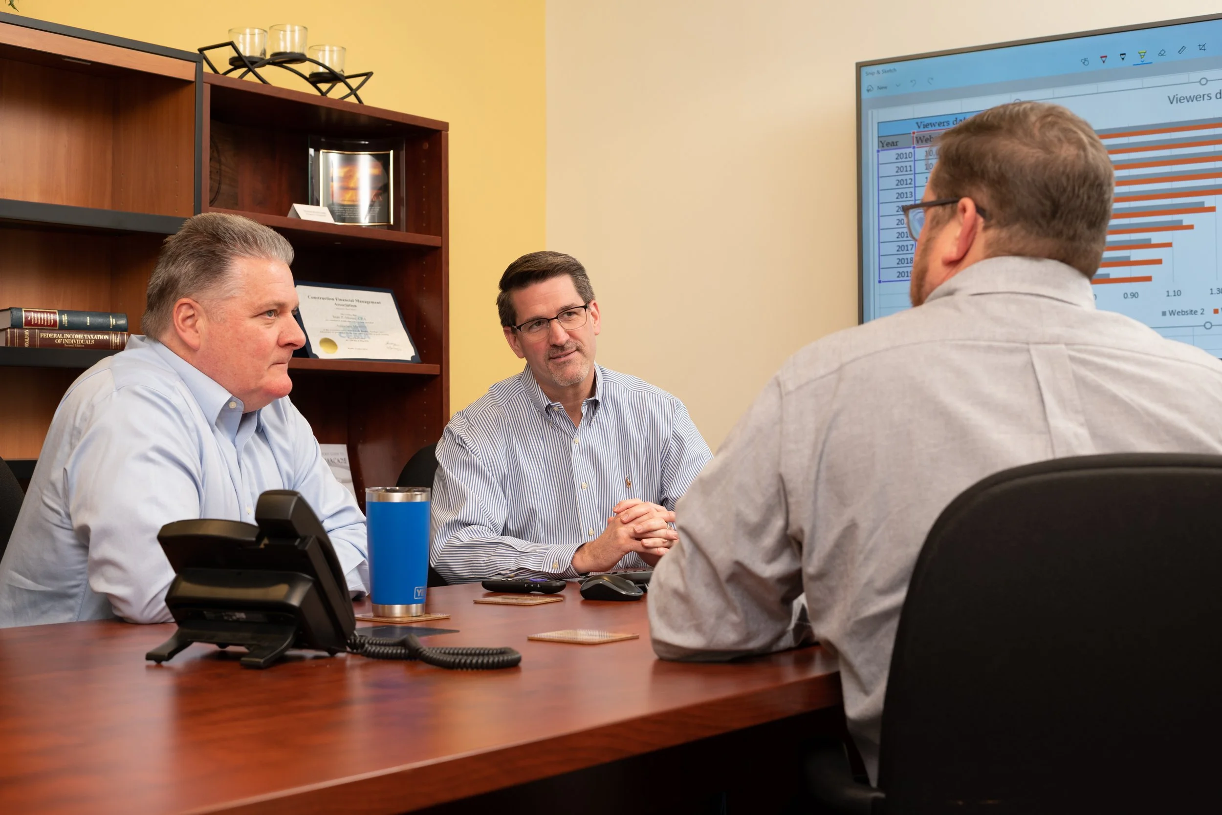 Three men in a business meeting in a conference room, with a large screen displaying a bar chart in the background.