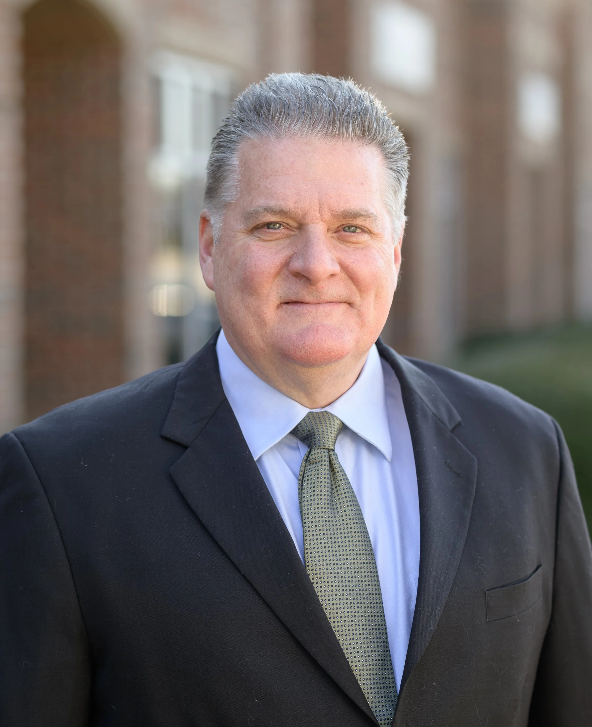 A middle-aged man in a suit standing outdoors in front of a brick building, looking at the camera with a slight smile.