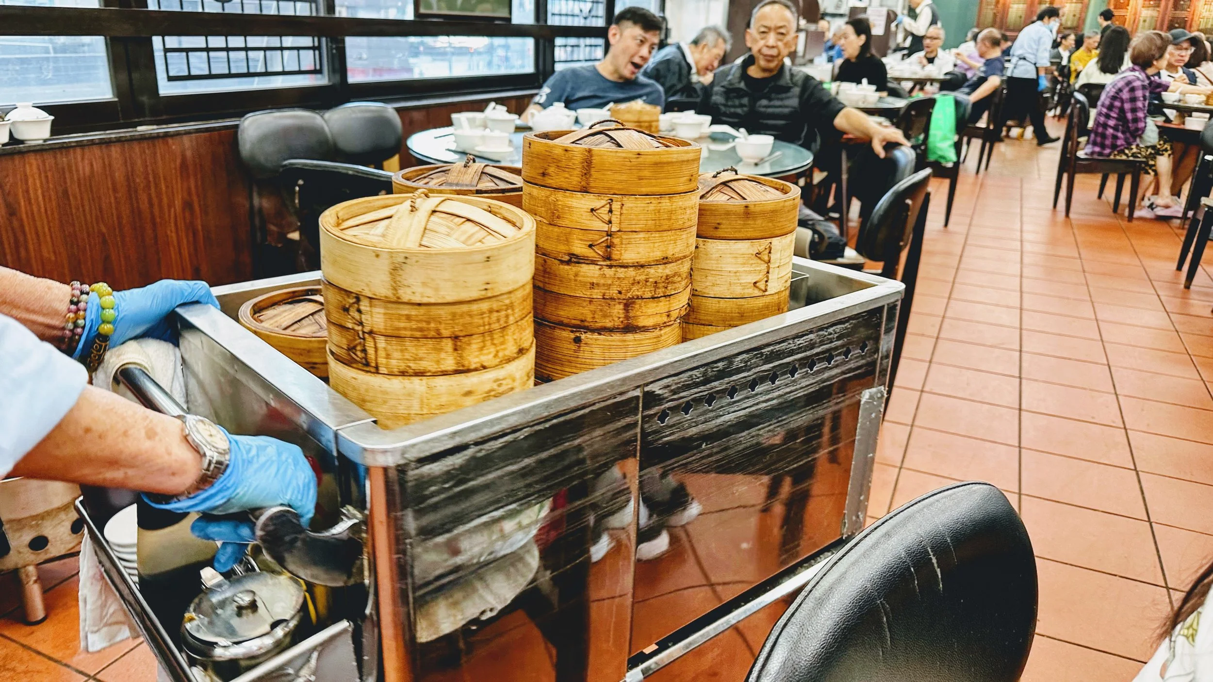 A food cart with stacked bamboo steamers in a busy Asian restaurant. Diners are seated at tables in the background.