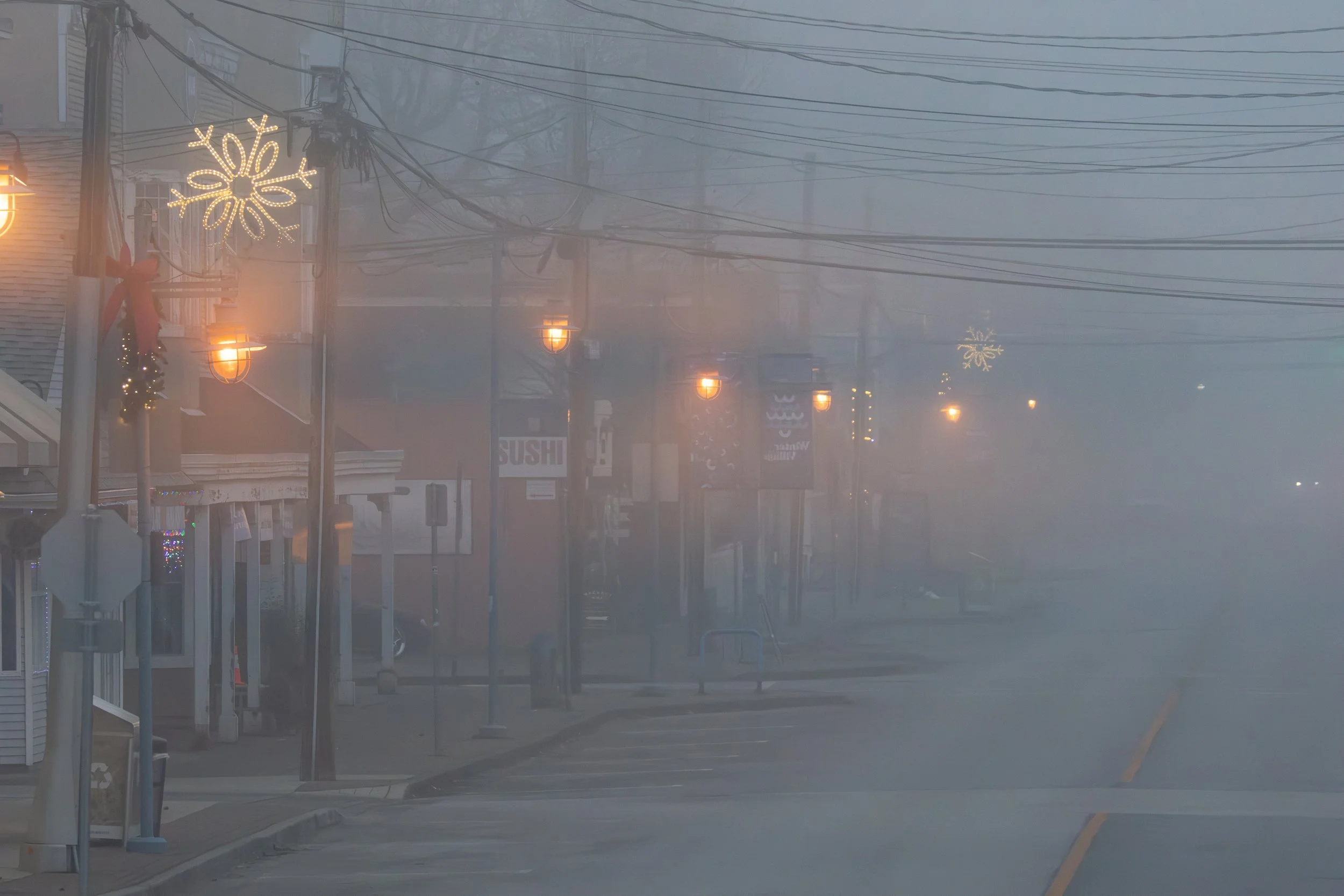 A foggy street scene with decorative holiday lights hanging from poles, including snowflake and star shapes, and street lamps. Buildings with signs, including one labeled 'SUSHI,' are visible in the background.