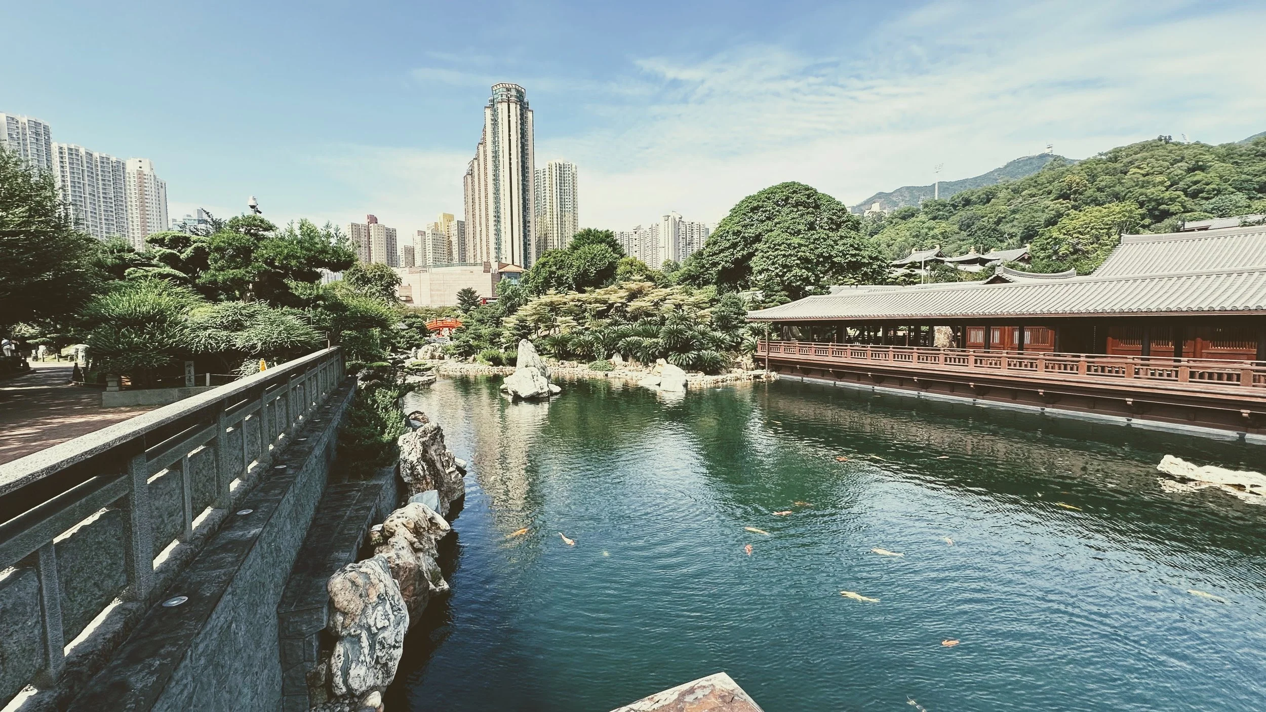 Traditional Asian building with wooden structure and gray tiled roof along a calm pond with koi fish, surrounded by lush greenery and tall modern skyscrapers in the background under a blue sky.