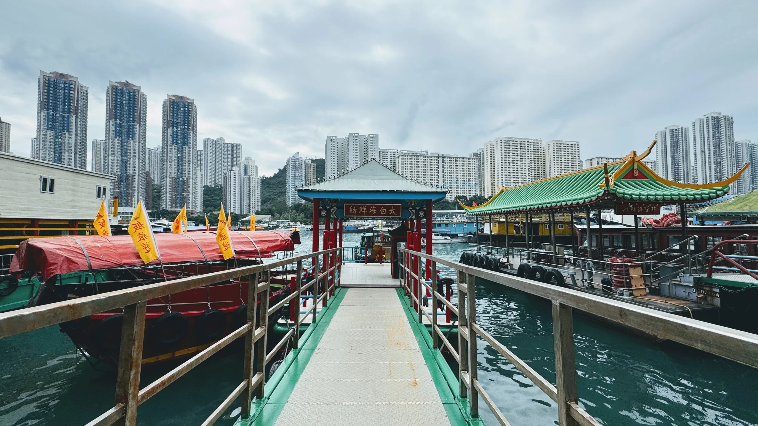 Dock leading to boats in harbor with high-rise buildings in background.