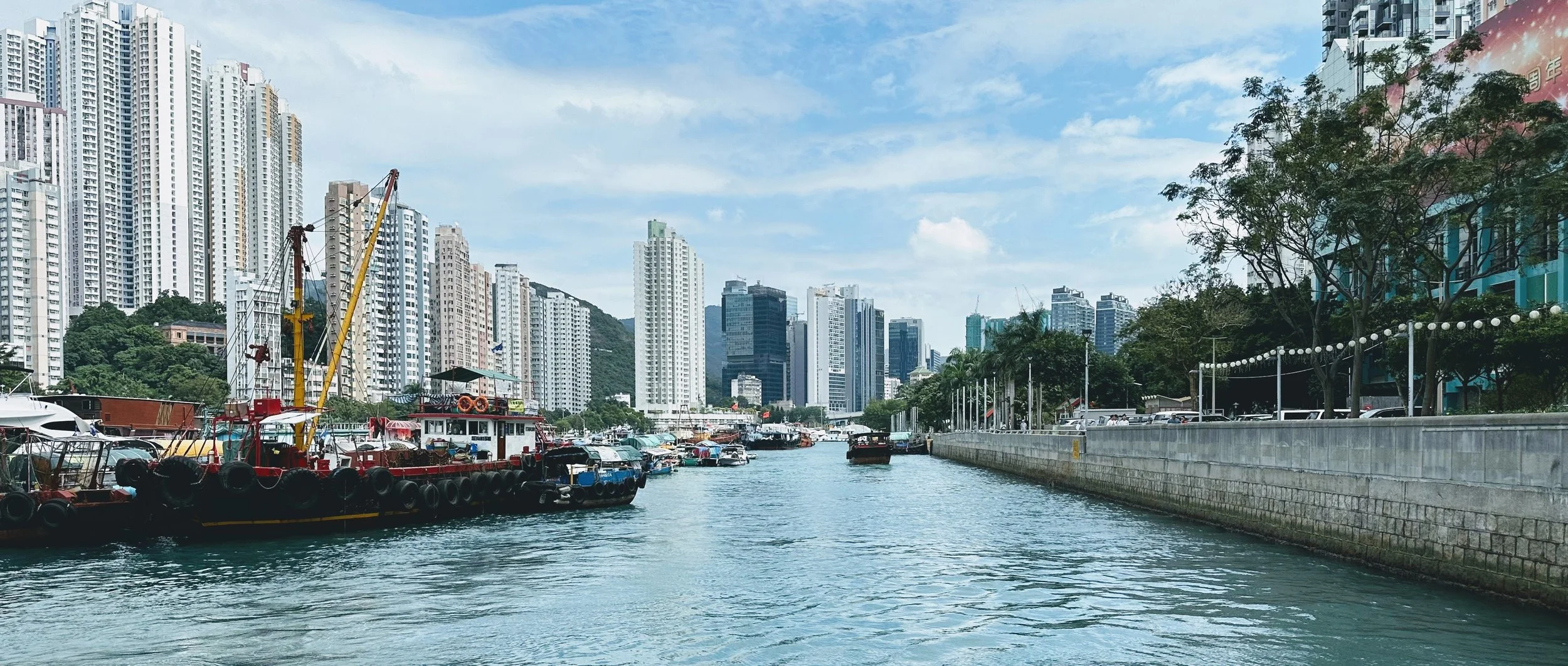 View of a harbor with boats and high-rise buildings in the background, trees lining the walkway along the water, clear blue skies with some clouds.