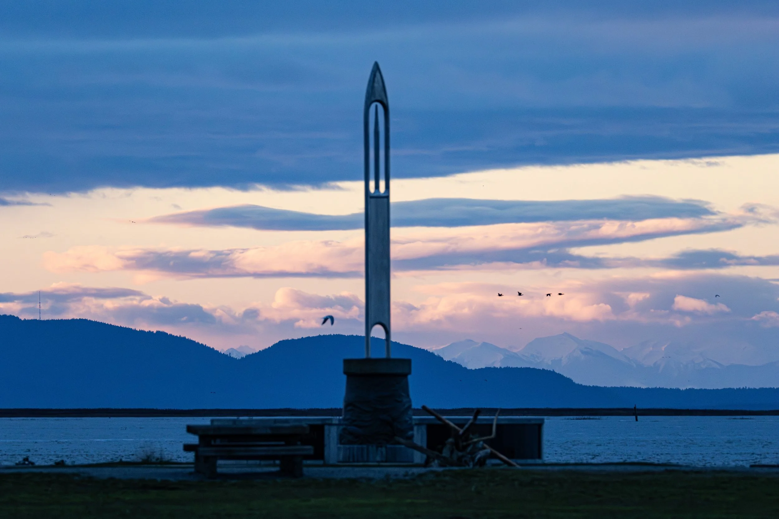 Large metal sculpture resembling a rocket, positioned on a platform near a body of water with mountains in the background and a cloudy sky.