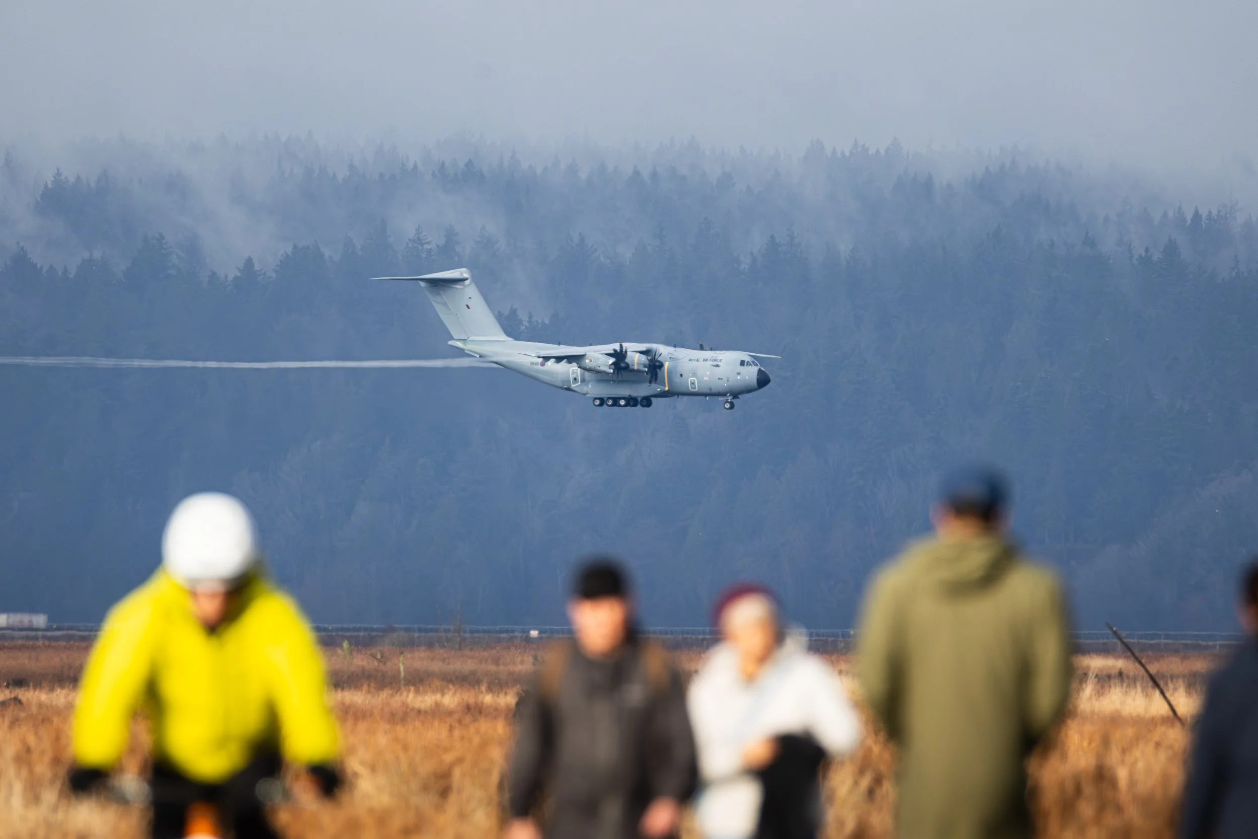 Military aircraft flying low over a field as a group of people watch from the ground, with a forest and mountains in the background.