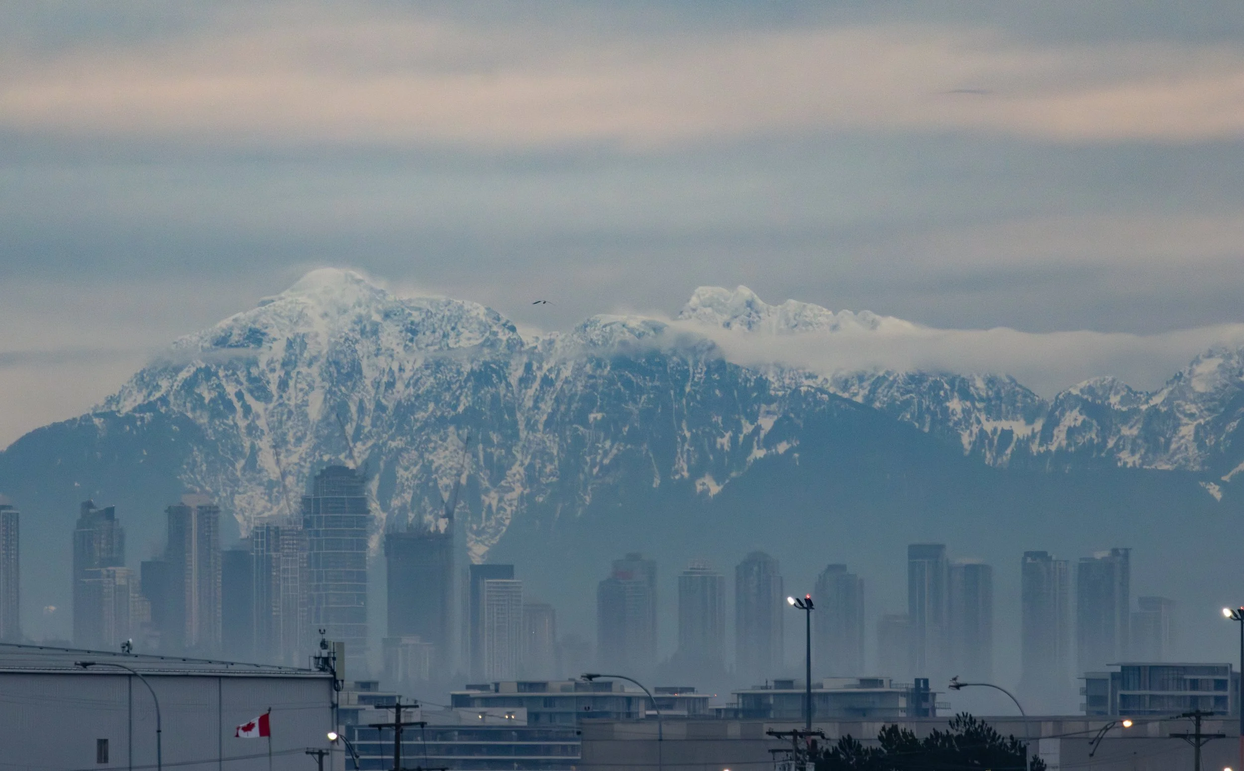 Cityscape with tall buildings in front of large snow-capped mountains and cloudy sky.