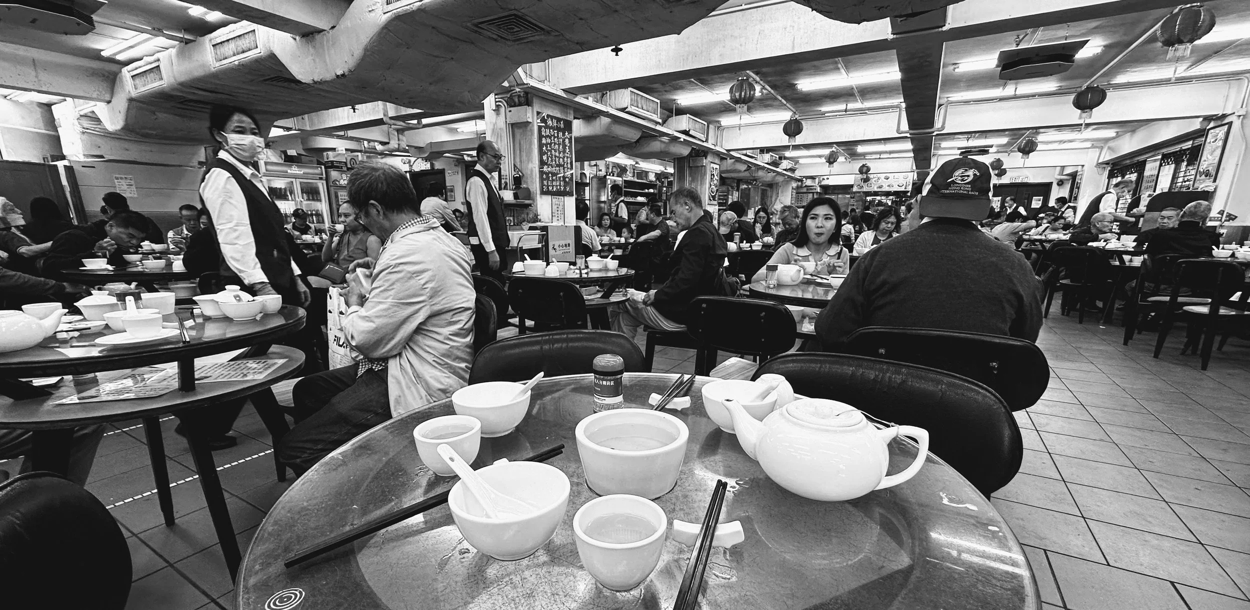 Black and white photo of a busy Asian restaurant with several patrons seated at tables, eating and chatting. The foreground shows an empty round table with bowls, a teapot, and chopsticks. Staff members are serving and attending to customers in the b