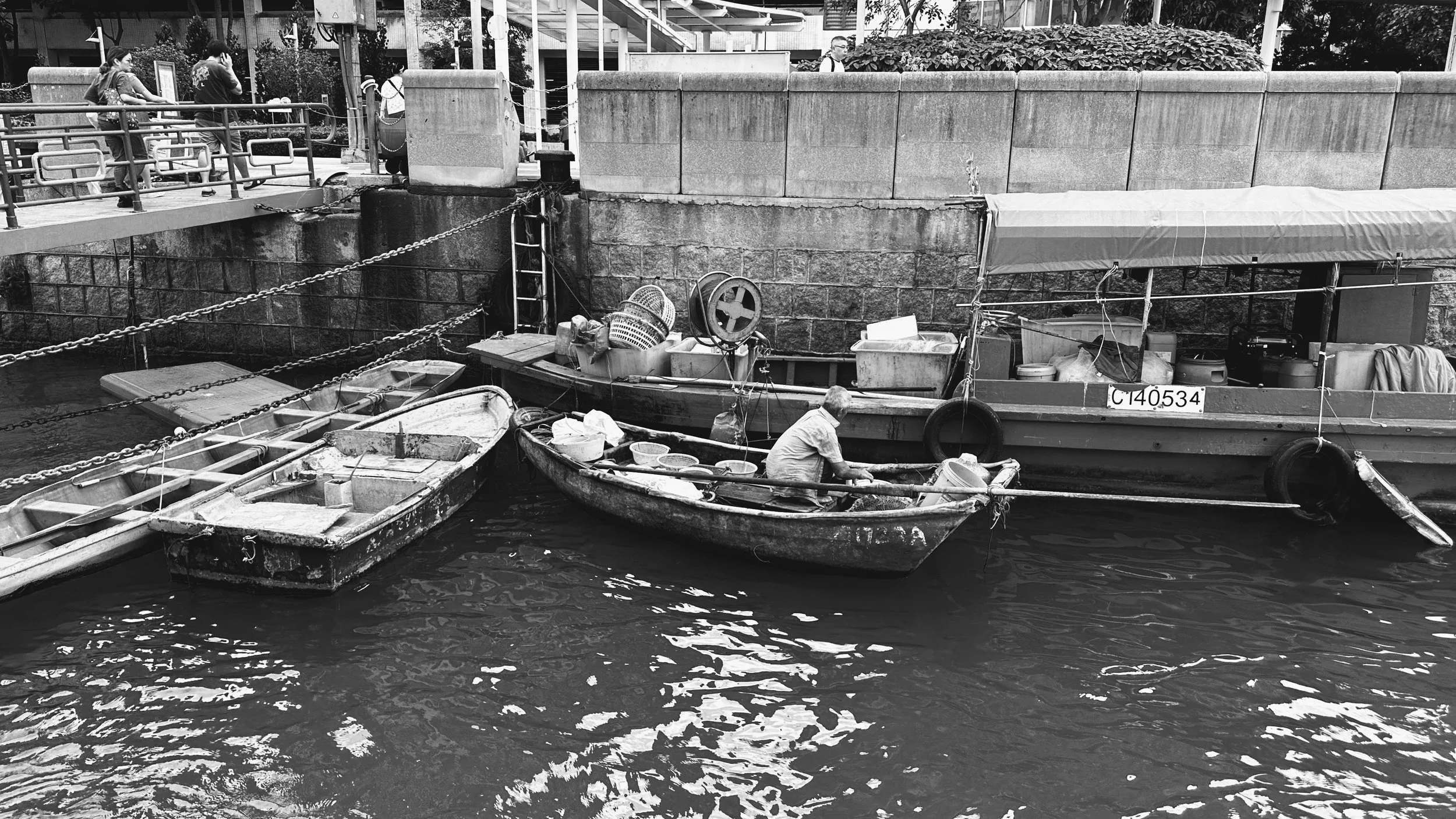 A small fishing boat docked next to a larger boat with a person sitting in it, surrounded by water and various equipment. Behind the boats, there is a stone wall and a railing, with several people walking and standing on the bridge.
