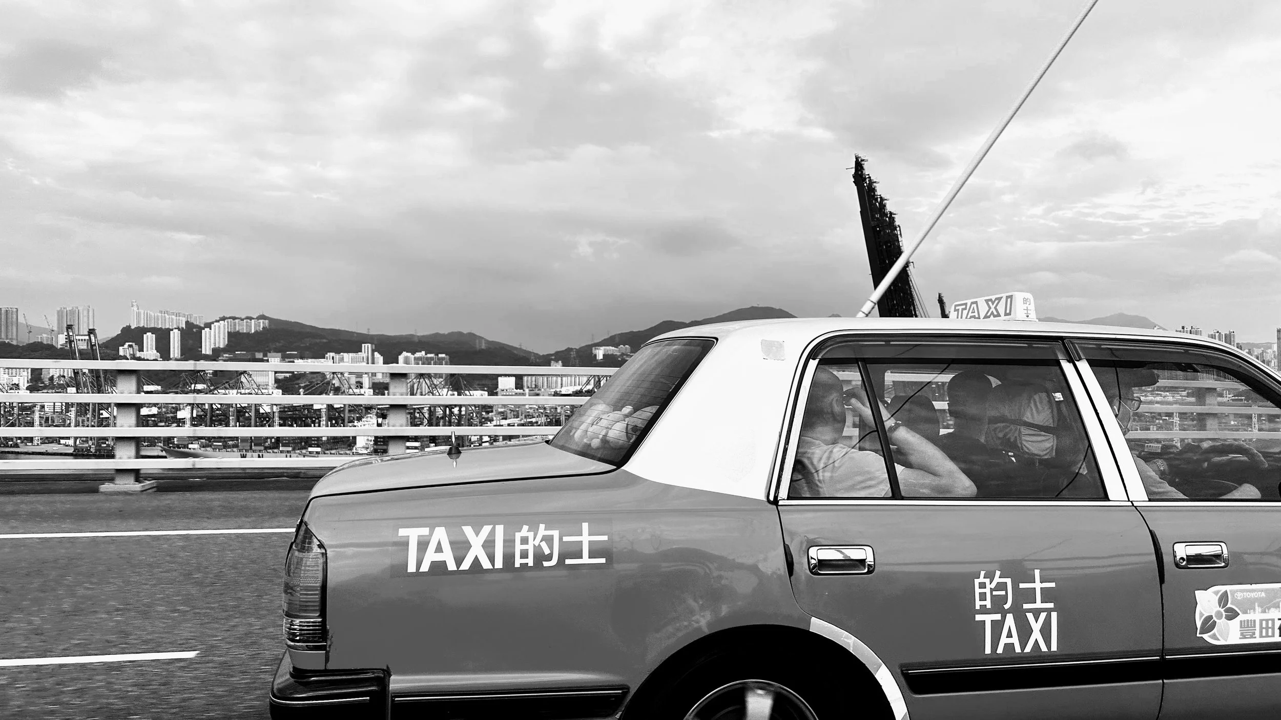 Black and white photo of a taxi on a bridge with mountains and city skyline in the background.