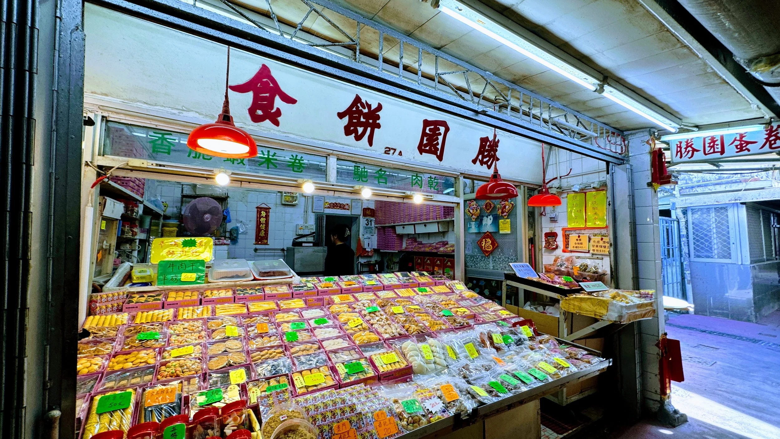 A traditional Asian market stall selling various dried goods and snacks, with red hanging lamps, a sign with Chinese characters, and a person working behind the counter.