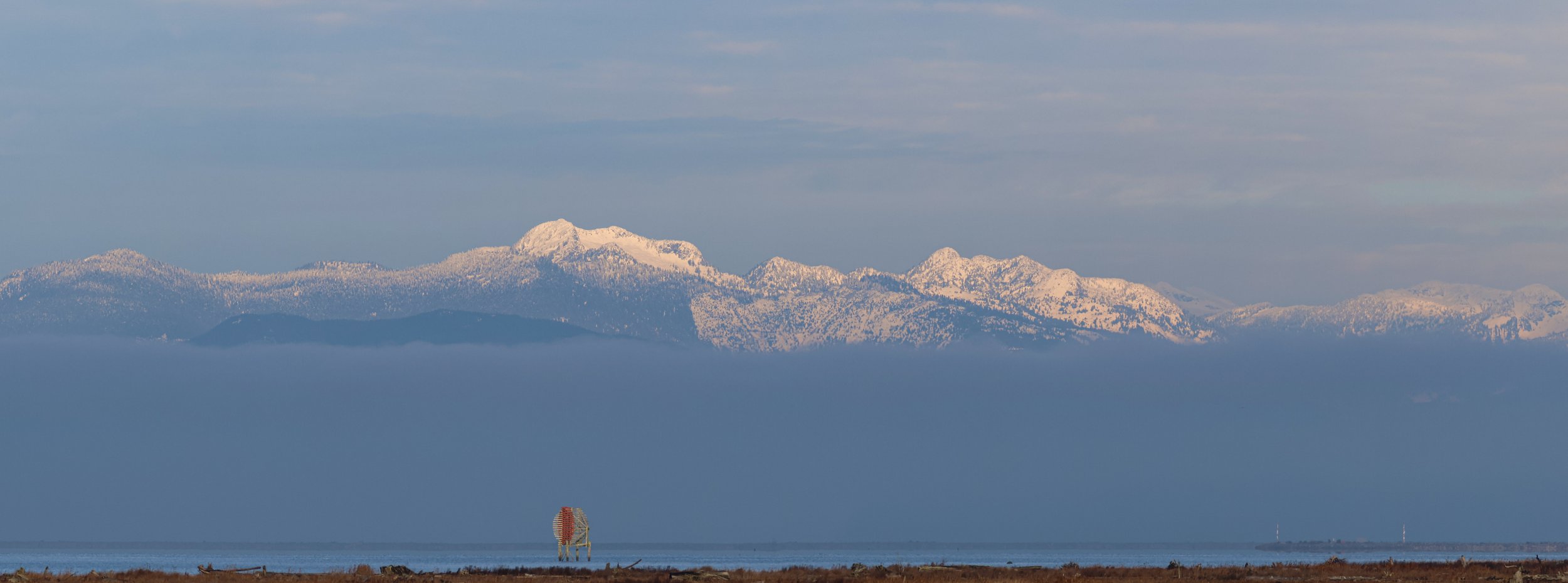 Snow-covered mountains in the distance above a body of water, with a small red and white navigational marker on the shoreline.