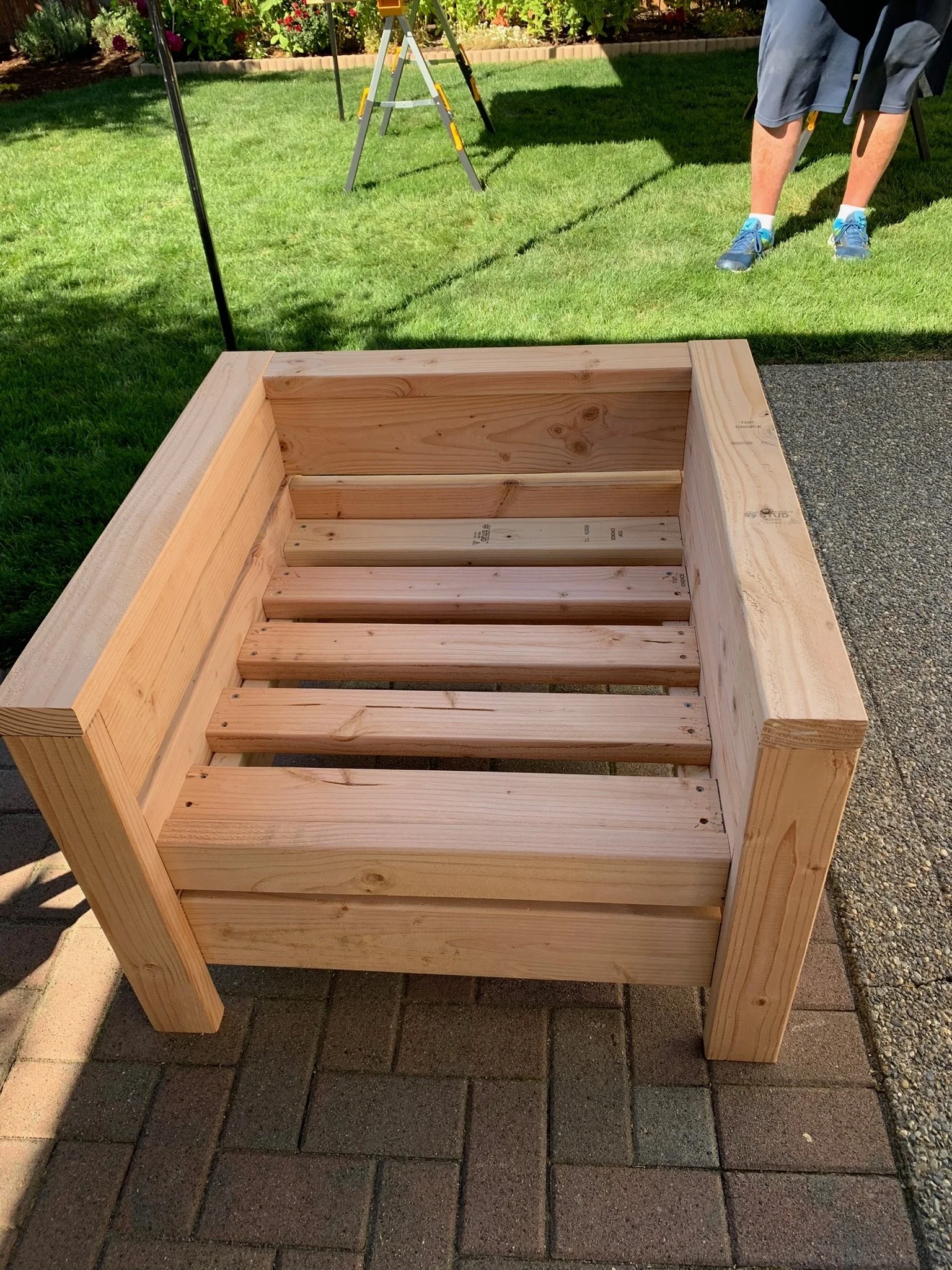 Unfinished wooden bed frame outdoors on brick and concrete, with grass and plants in the background.