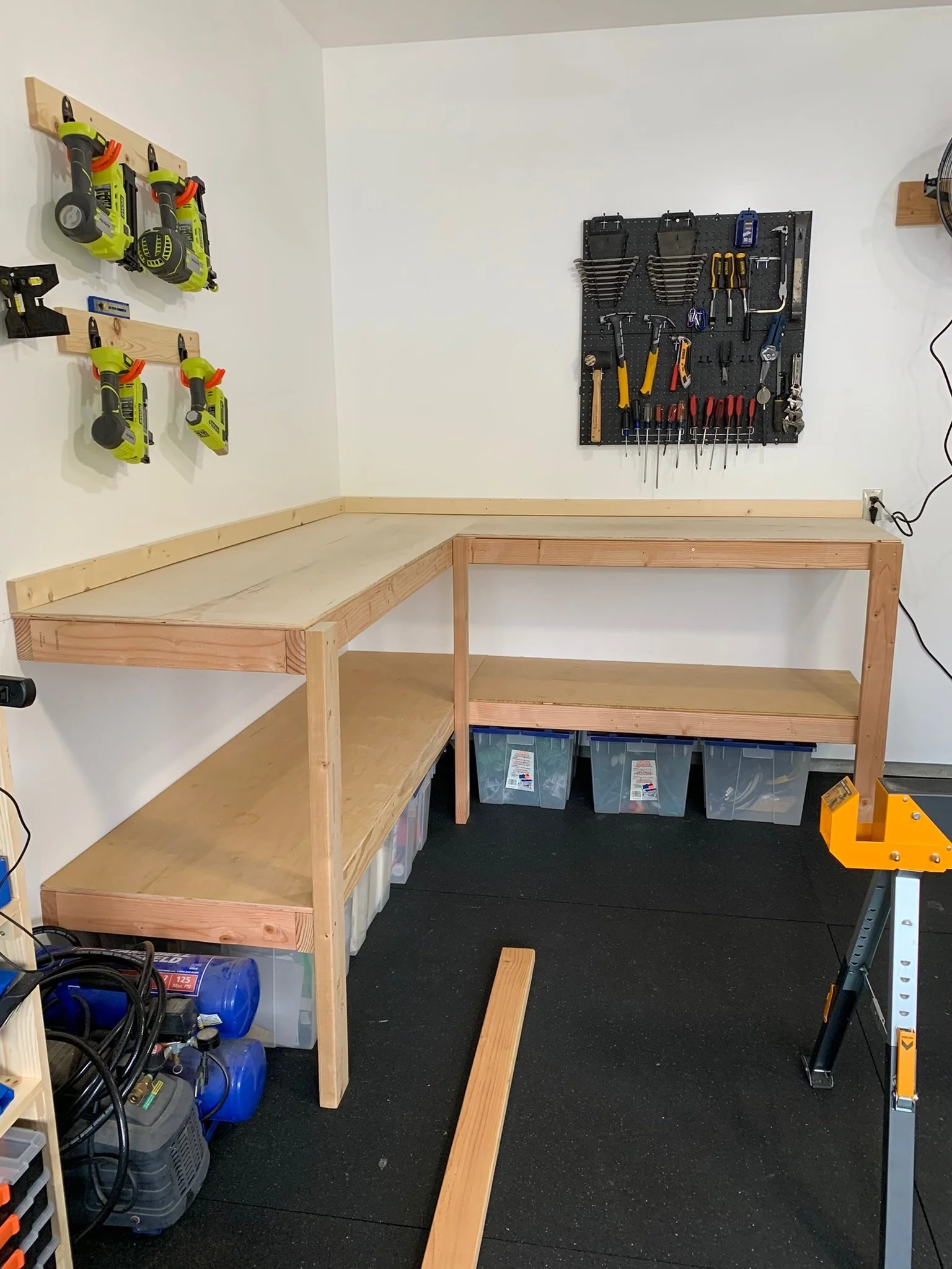 A woodworking workbench in a garage or workshop with tools and storage bins. The bench has a pegboard with assorted hand tools and a shelf with power tools on the wall.