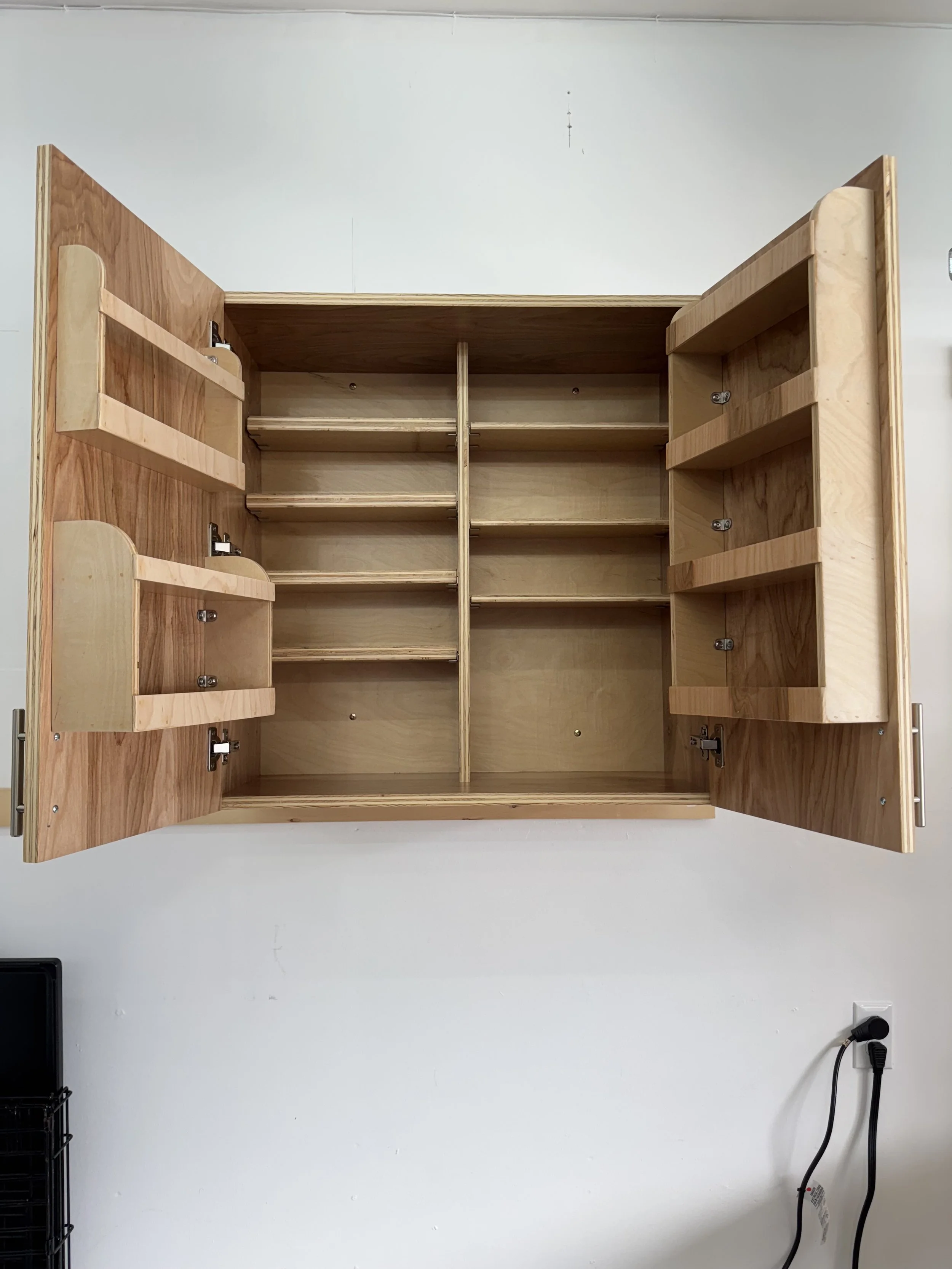 Empty unfinished wooden wall cabinet with open double doors, multiple shelves inside, mounted on a white wall near an electrical outlet.