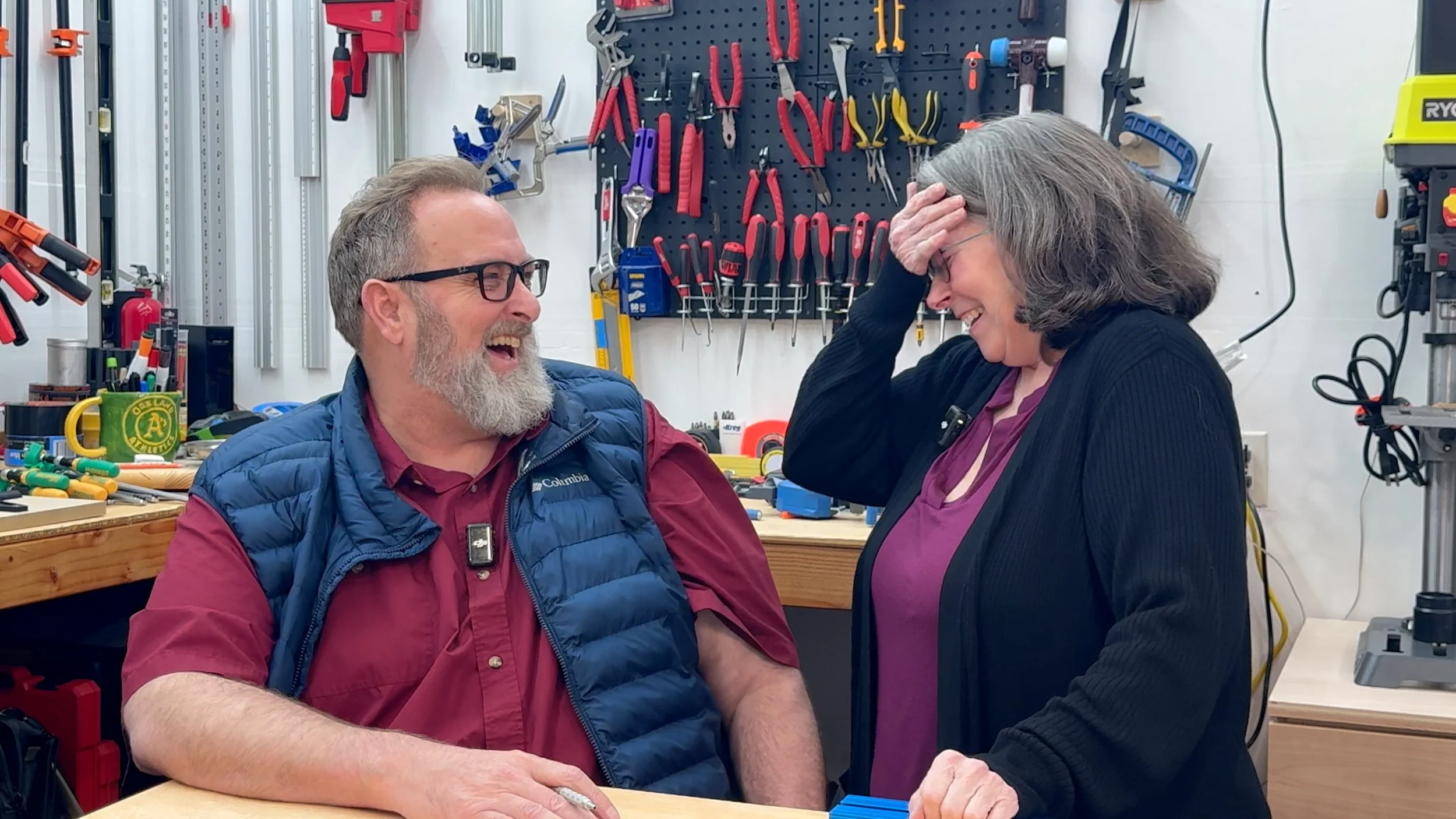 Two people, a man and a woman, are laughing in a workshop. The man has a beard, glasses, and is wearing a blue vest over a maroon shirt. The woman has gray hair and is wearing a black sweater over a purple shirt. There are tools hanging on the wall behind them.