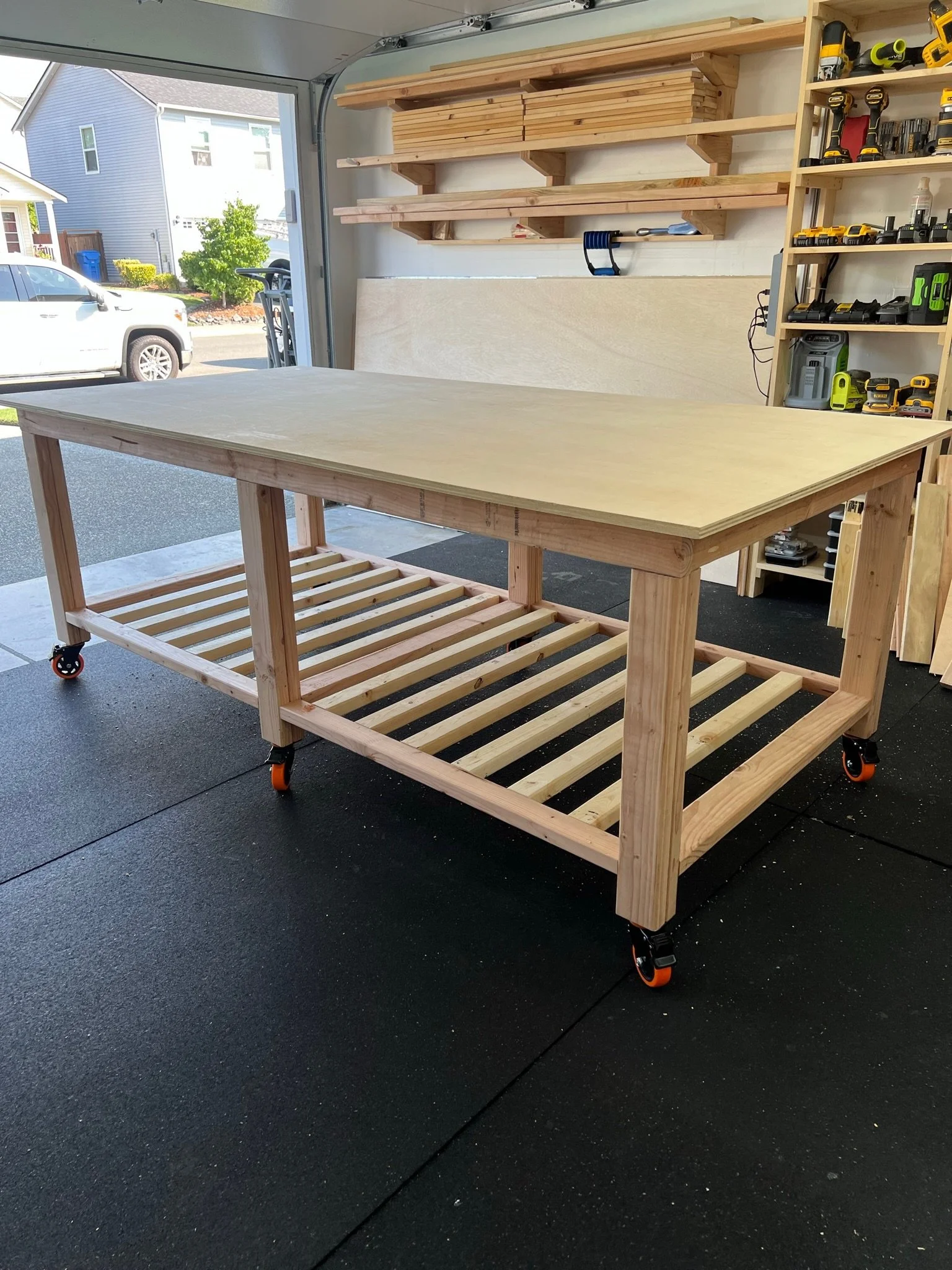 A woodworking workbench with a large plywood top and a slatted lower shelf, equipped with caster wheels, inside a cluttered garage with tools and wooden shelves.