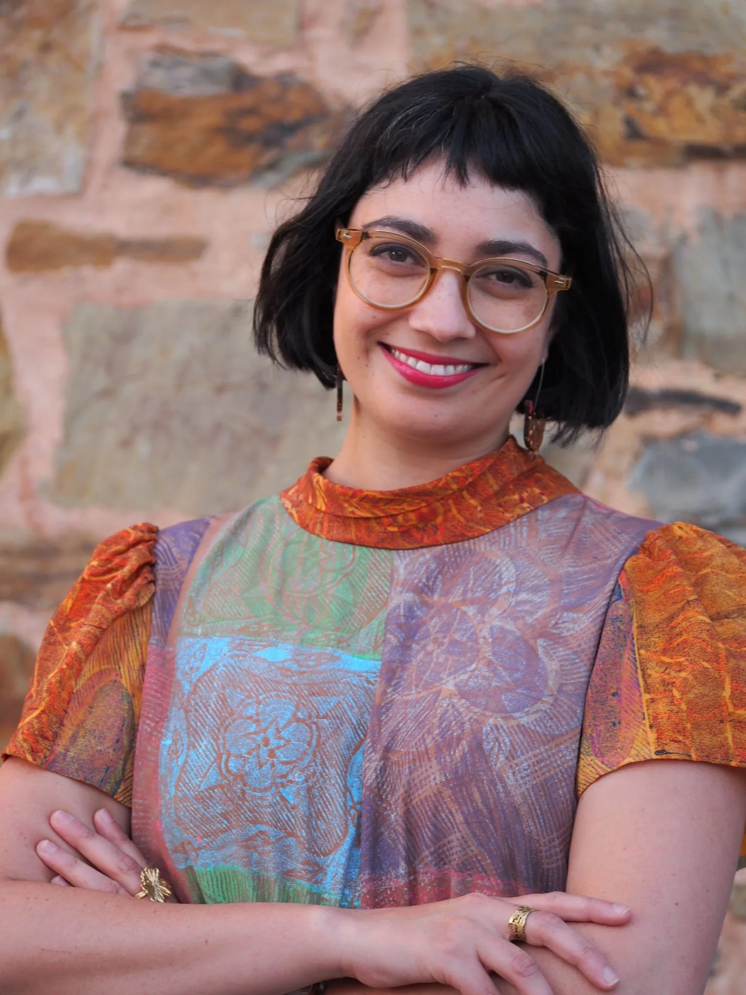 Close-up of a woman with short black hair, wearing glasses and colorful patterned clothing, smiling in front of a stone wall.