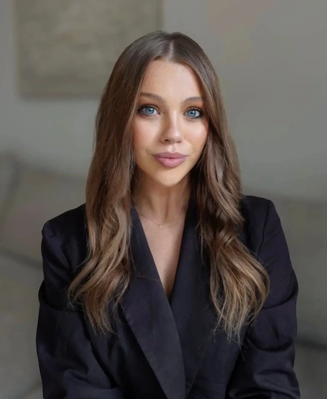 A woman with long, wavy brown hair and blue eyes, wearing a black blazer, sitting indoors with a neutral background.