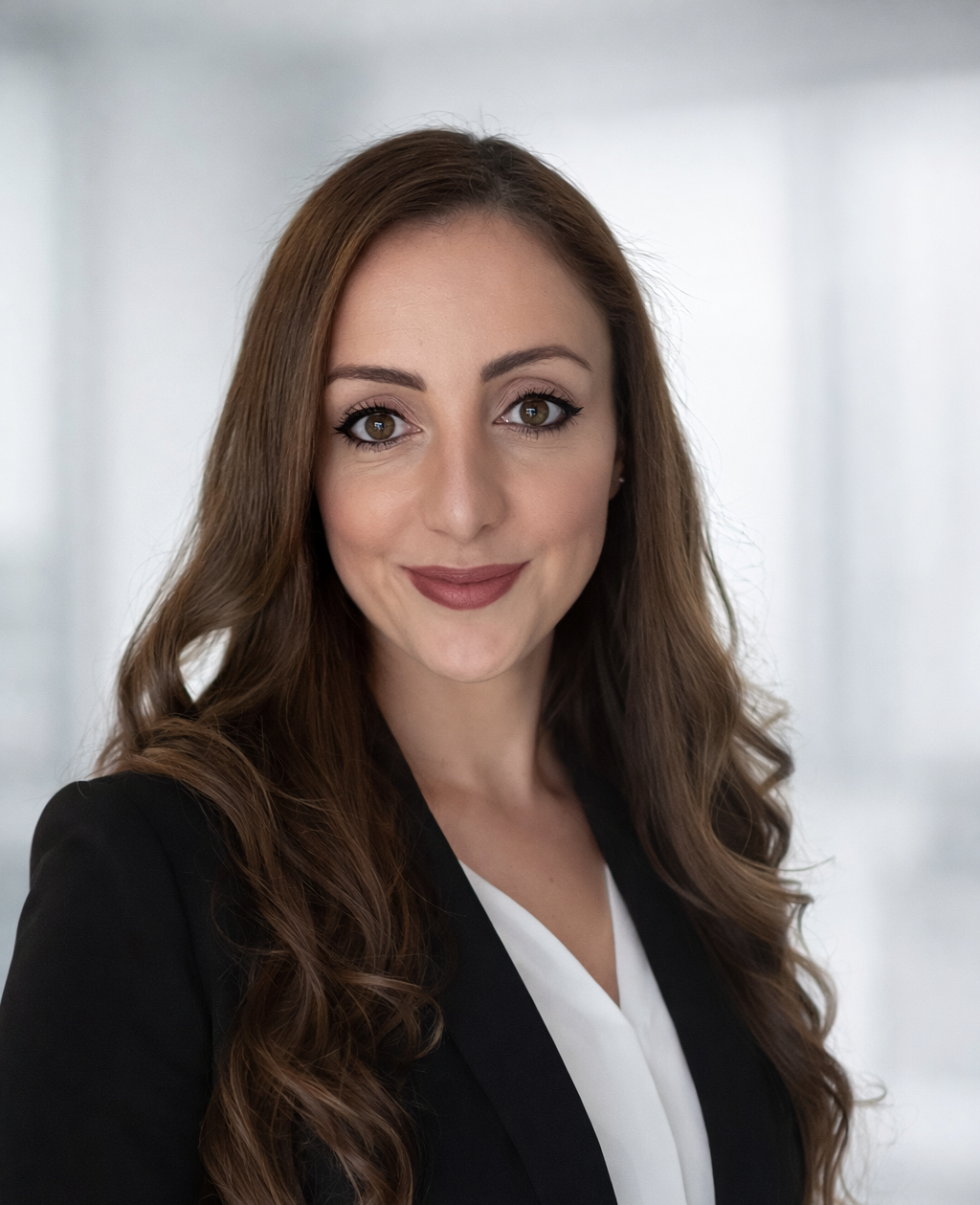 A woman with long brown hair, wearing a black blazer and white top, smiling at the camera.
