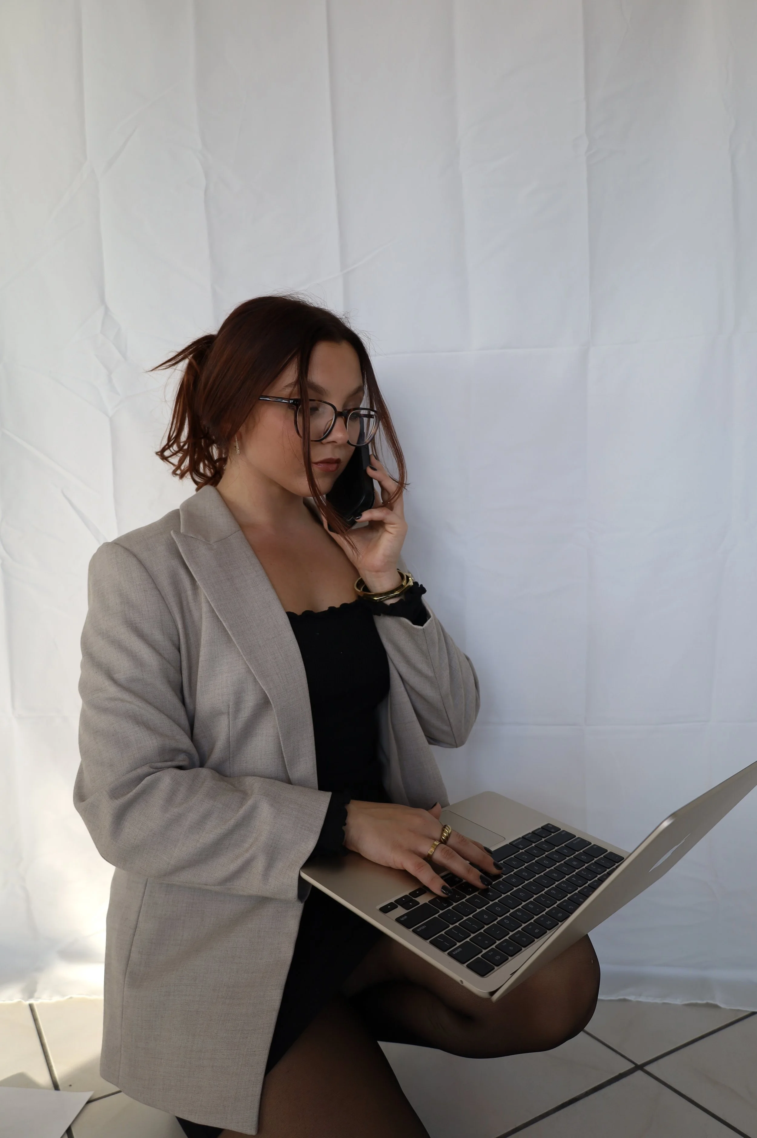A woman with shoulder-length brown hair, glasses, and wearing a beige blazer, is seated with her legs crossed, working on a laptop and talking on the phone against a white backdrop.