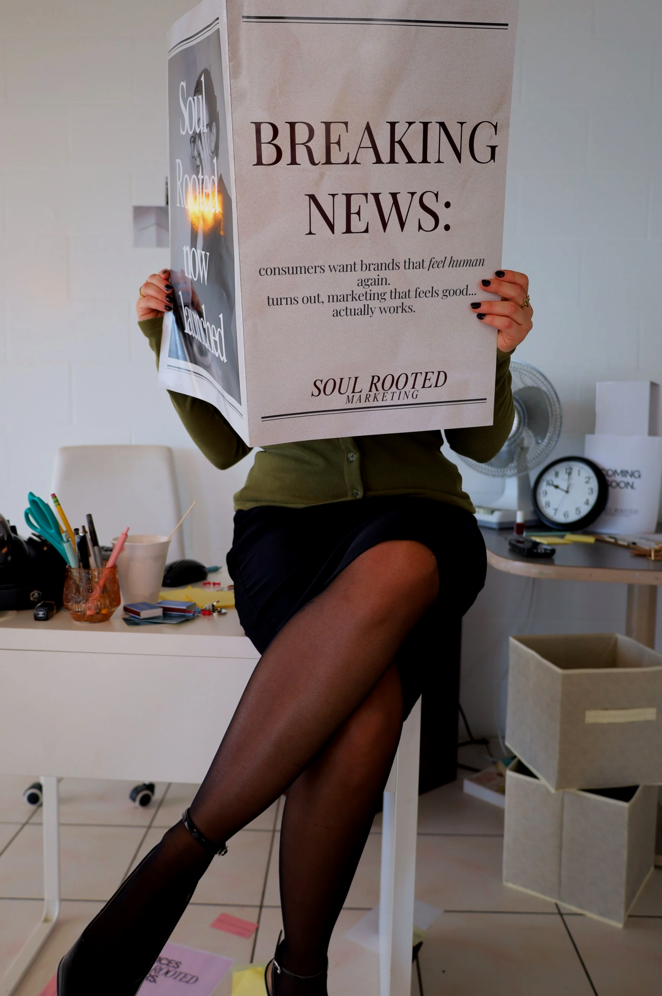 A woman sitting on a desk holding a large sign that reads "Breaking News: consumers want brands that feel human again" with the logo "Soul Rooted Marketing" at the bottom, in an office setting.