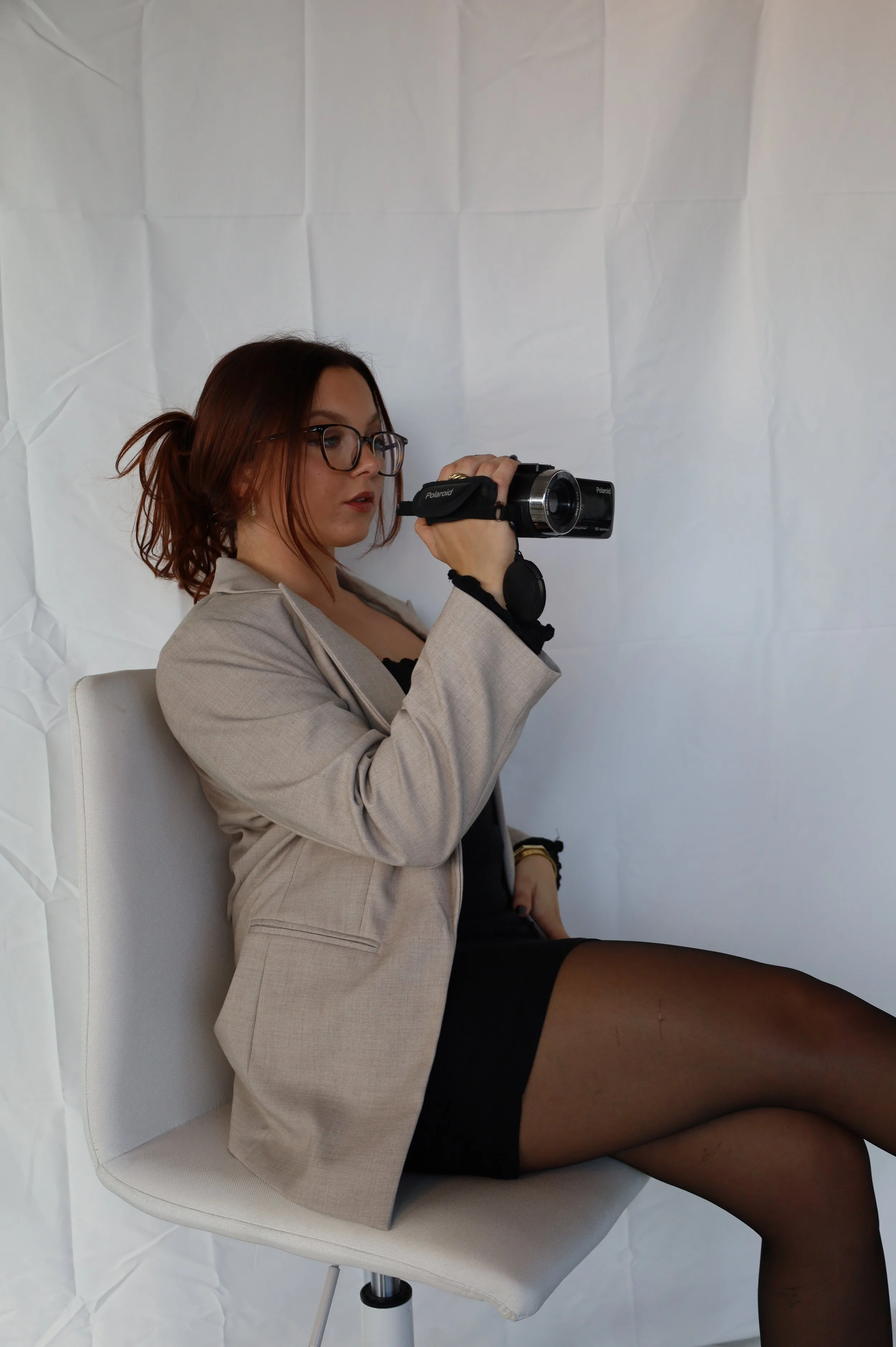 A woman with glasses and brown hair tied back sits on a white chair, holding a camera and looking to the side. She is wearing a beige blazer, black dress, and black tights, with a crumpled white background behind her.