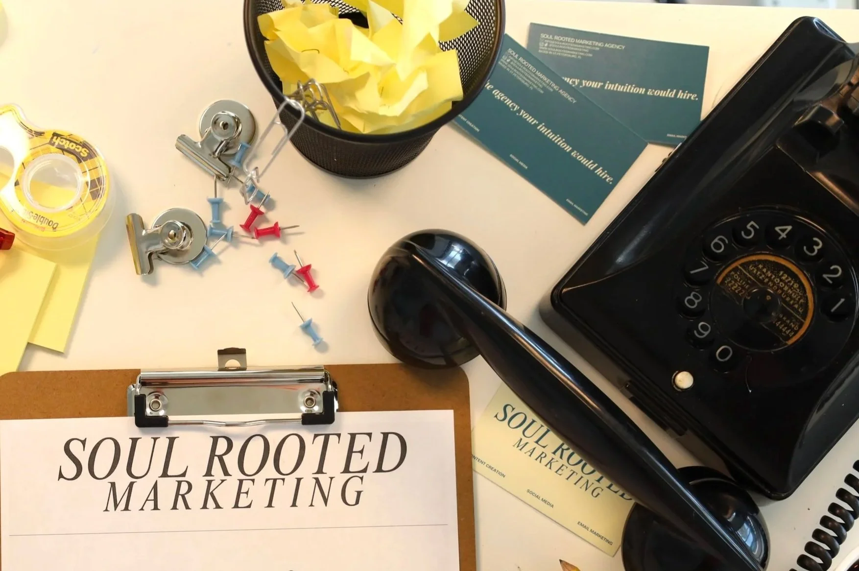 A cluttered office desk with a business card labeled 'Soul Rooted Marketing,' a black vintage rotary phone, a clipboard with the same company name, a yellow tape dispenser, colored push pins, a black mesh pen holder filled with yellow tissue paper, business flyers, and a black pen.