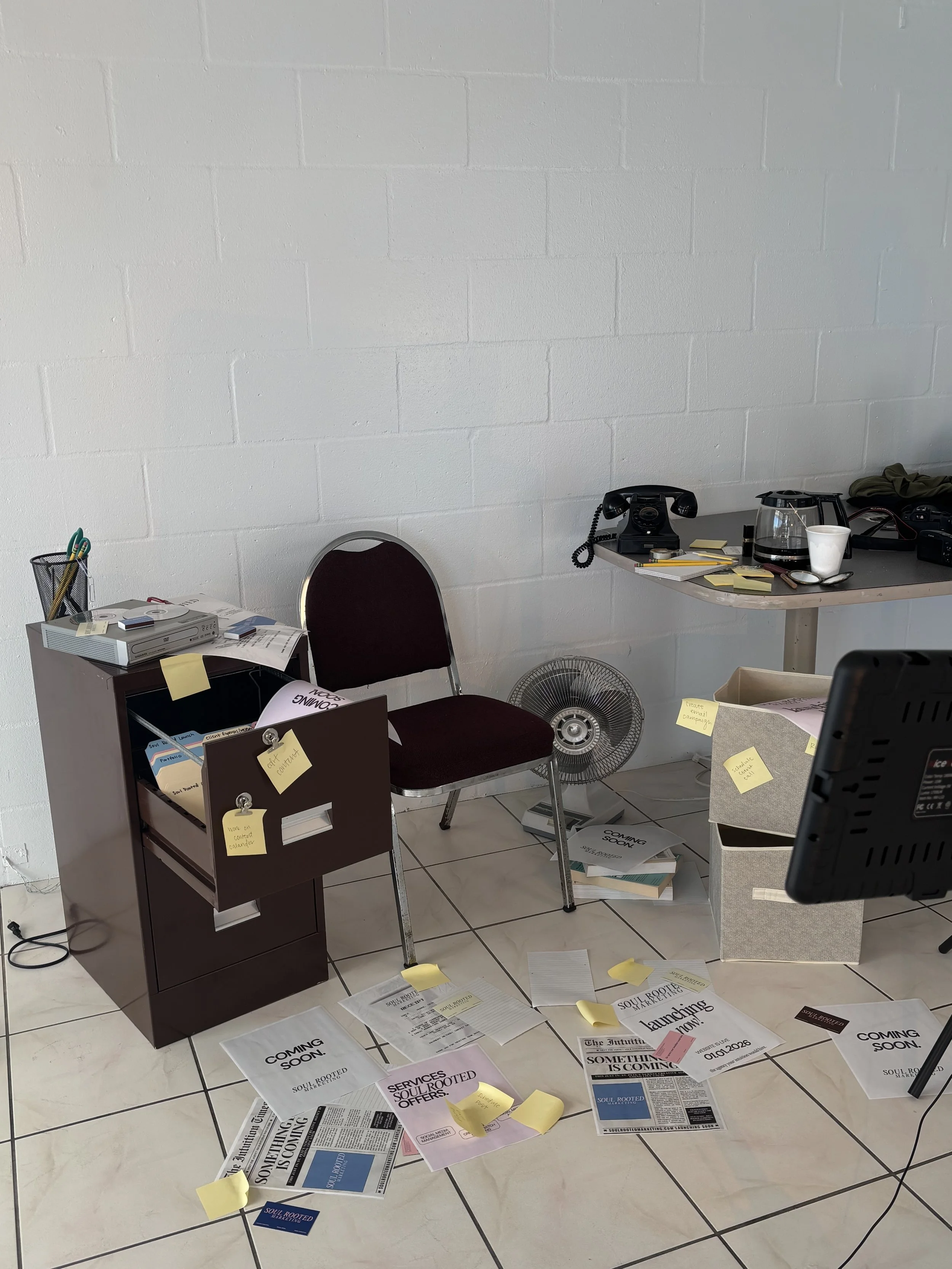Office desk area with scattered papers, sticky notes, a vintage black rotary phone, a glass of water, and a fan. A maroon chair sits in front of a small filing cabinet with papers and sticky notes. The floor is tiled and the background wall is plain white.