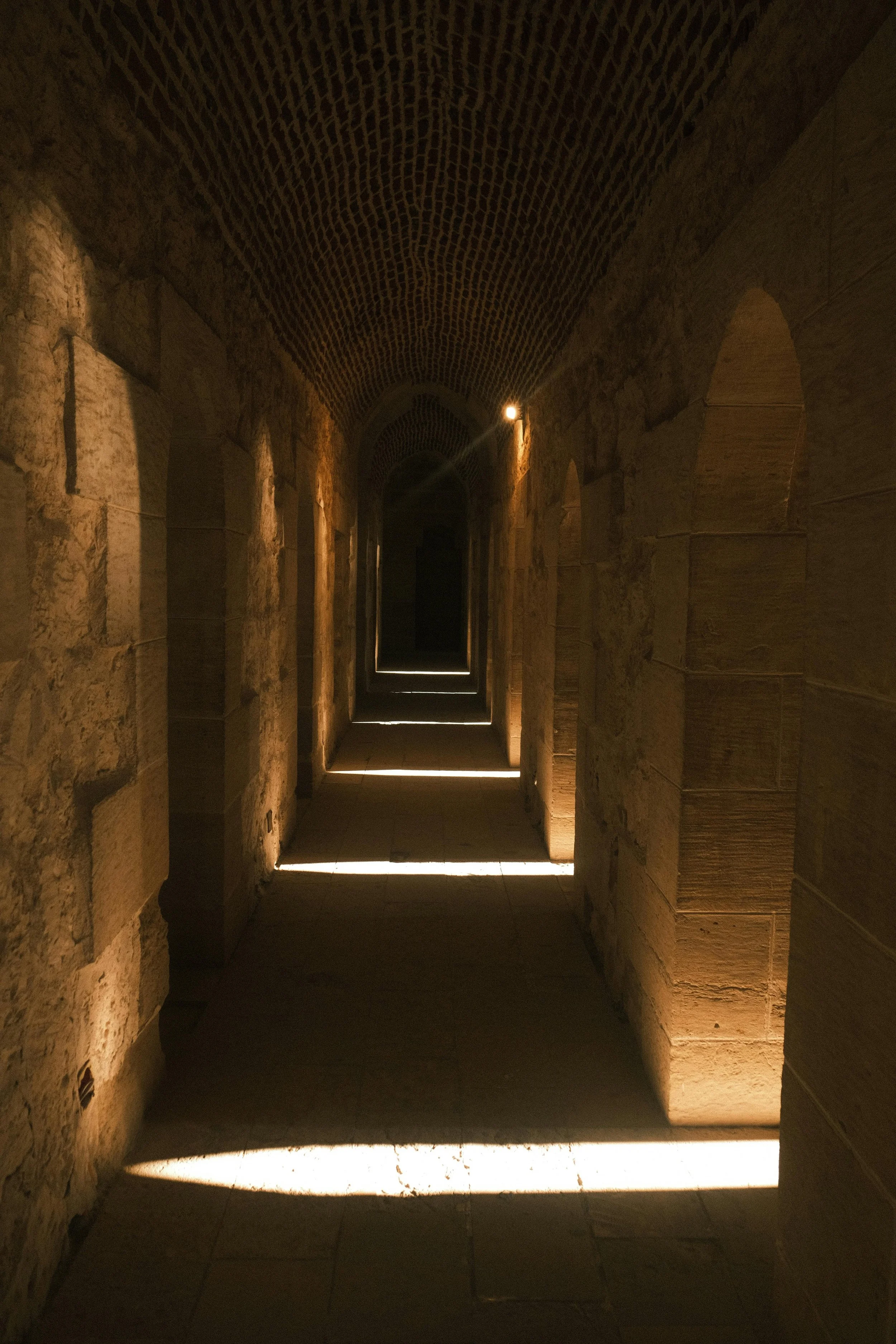 A dimly lit stone corridor with arched openings on the right side and light strips on the floor.